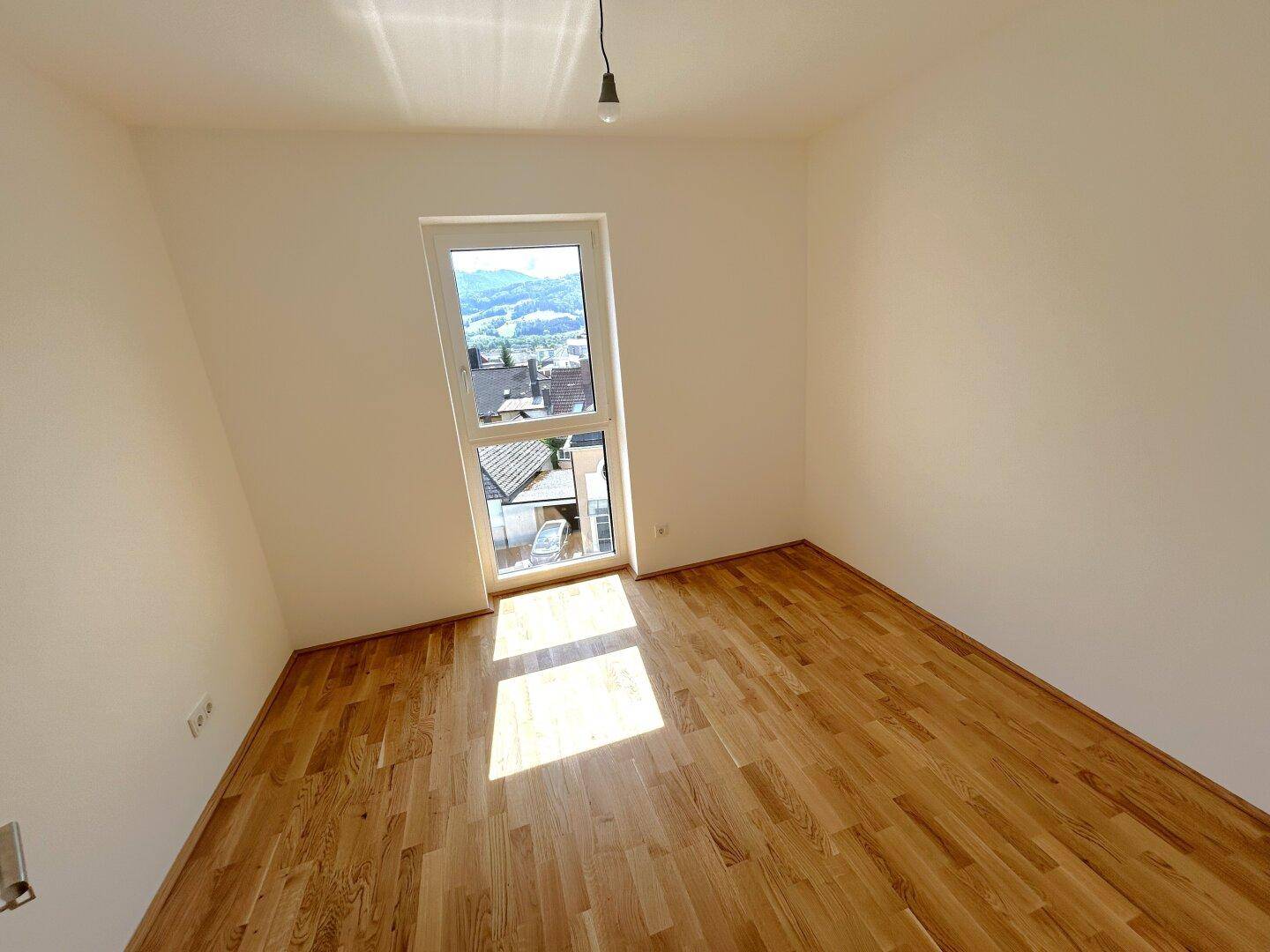 Helles Schlafzimmer mit Parkettboden und einem Fenster mit Blick auf die Umgebung.