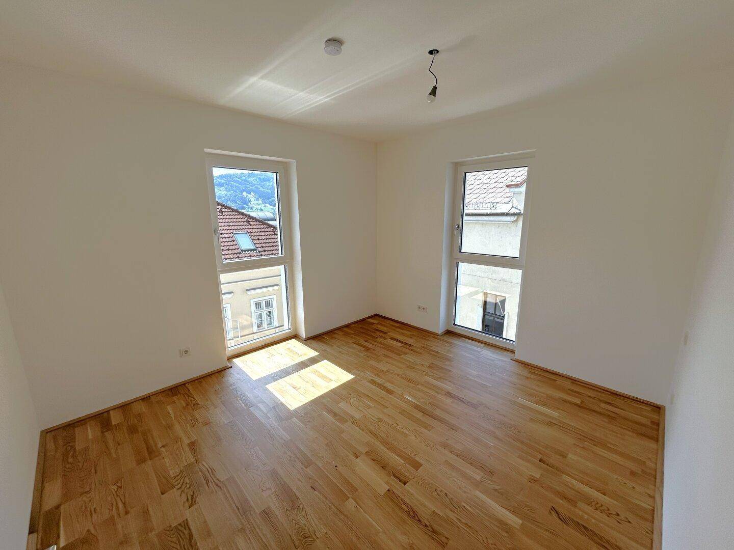 Helles Schlafzimmer mit Parkettboden und zwei Fenstern, die einen Blick ins Grüne ermöglichen.