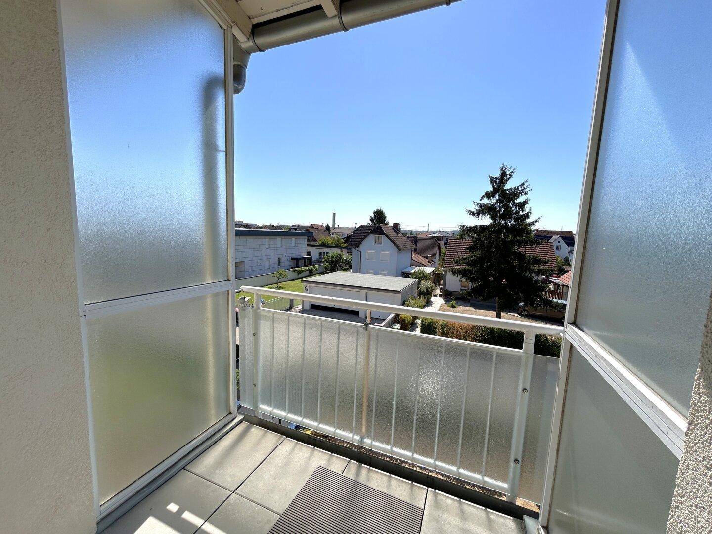 Heller Balkon mit weitem Blick über die umliegende Wohngegend und viel Grün unter klarem Himmel.