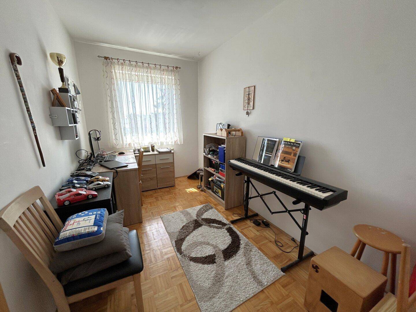 Arbeitszimmer mit Laminatboden, Schreibtisch und Musikinstrumenten am Fenster, gut beleuchtet.
