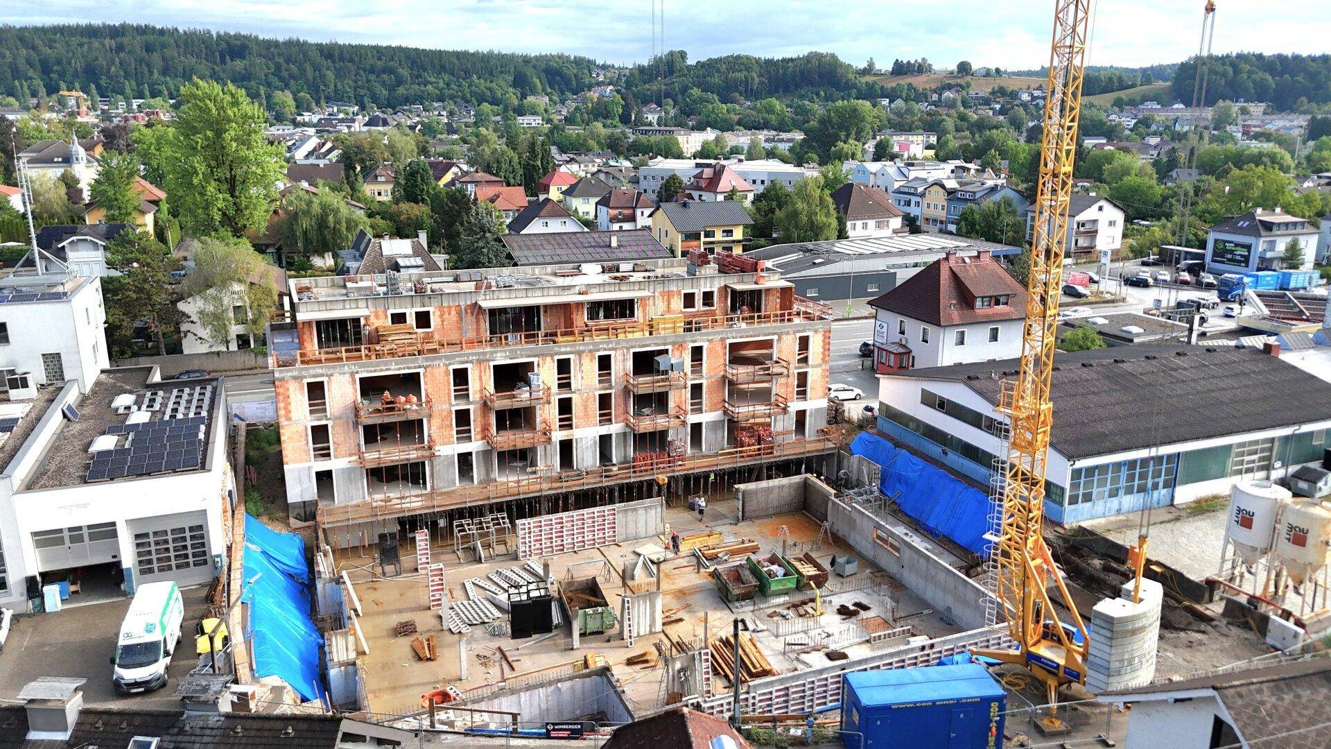 Drohnenansicht eines Neubaus mit Baugrube und umliegender Wohnbebauung in grüner Umgebung.
