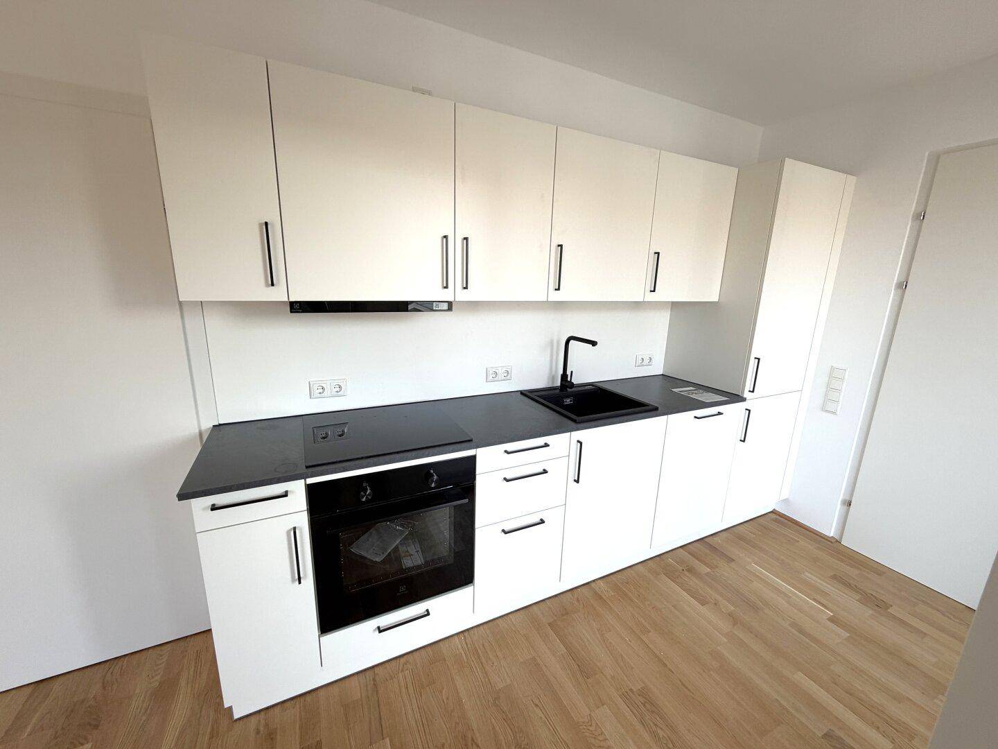 Moderne Einbauküche mit weißen Schränken, schwarzer Arbeitsplatte und integriertem Backofen auf hellem Holzfußboden.