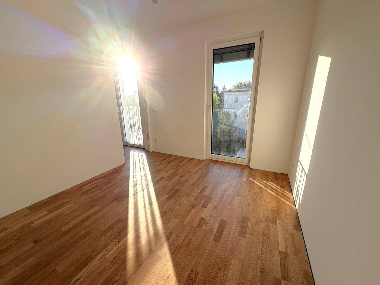 Lichtdurchflutetes Zimmer mit Holzboden und großen Fenstern, die viel Sonnenlicht hereinlassen.