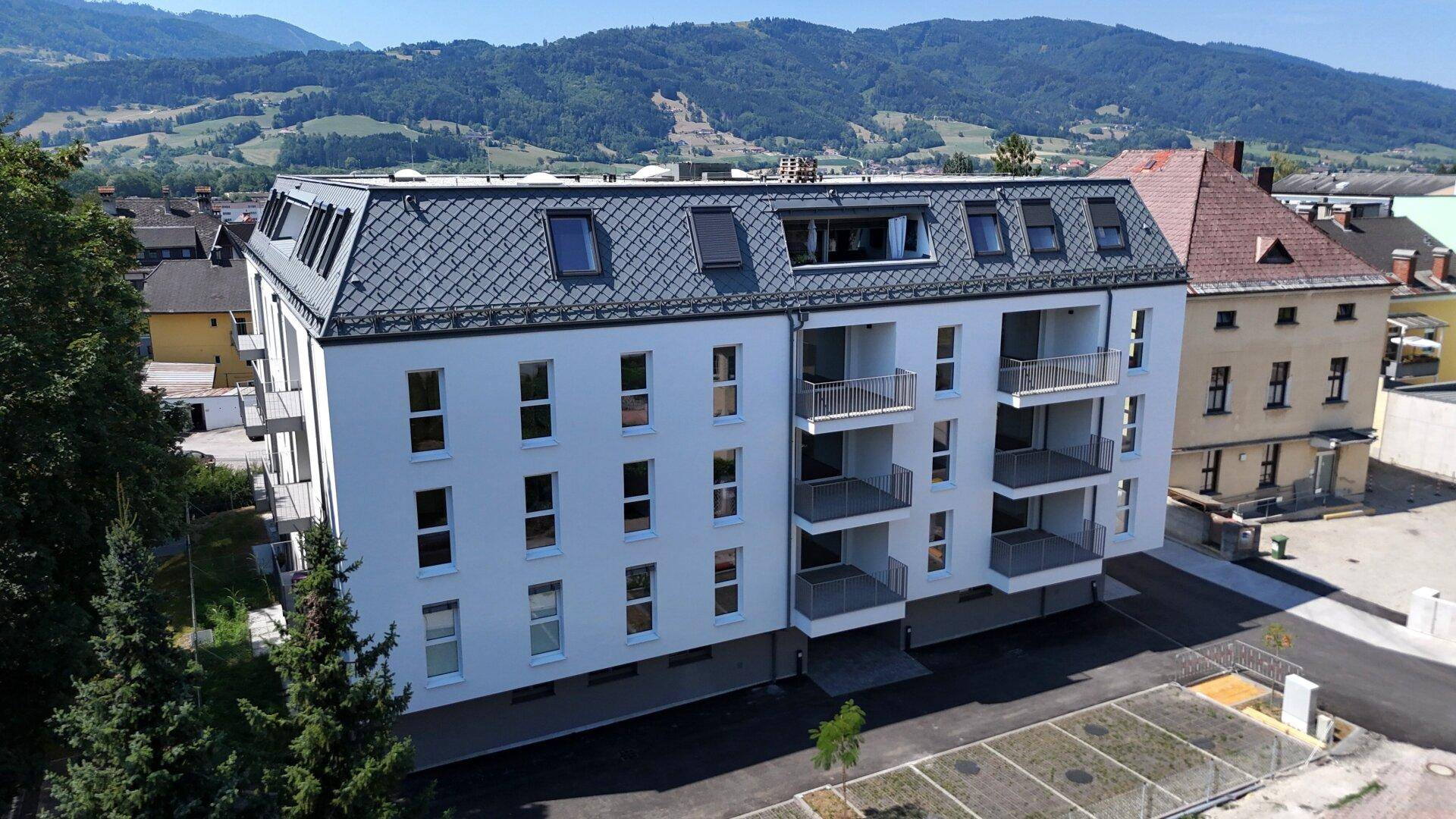 Neubau mit weißer Fassade, Balkonen und Dachfenstern, eingebettet in eine grüne Landschaft mit Bergen.