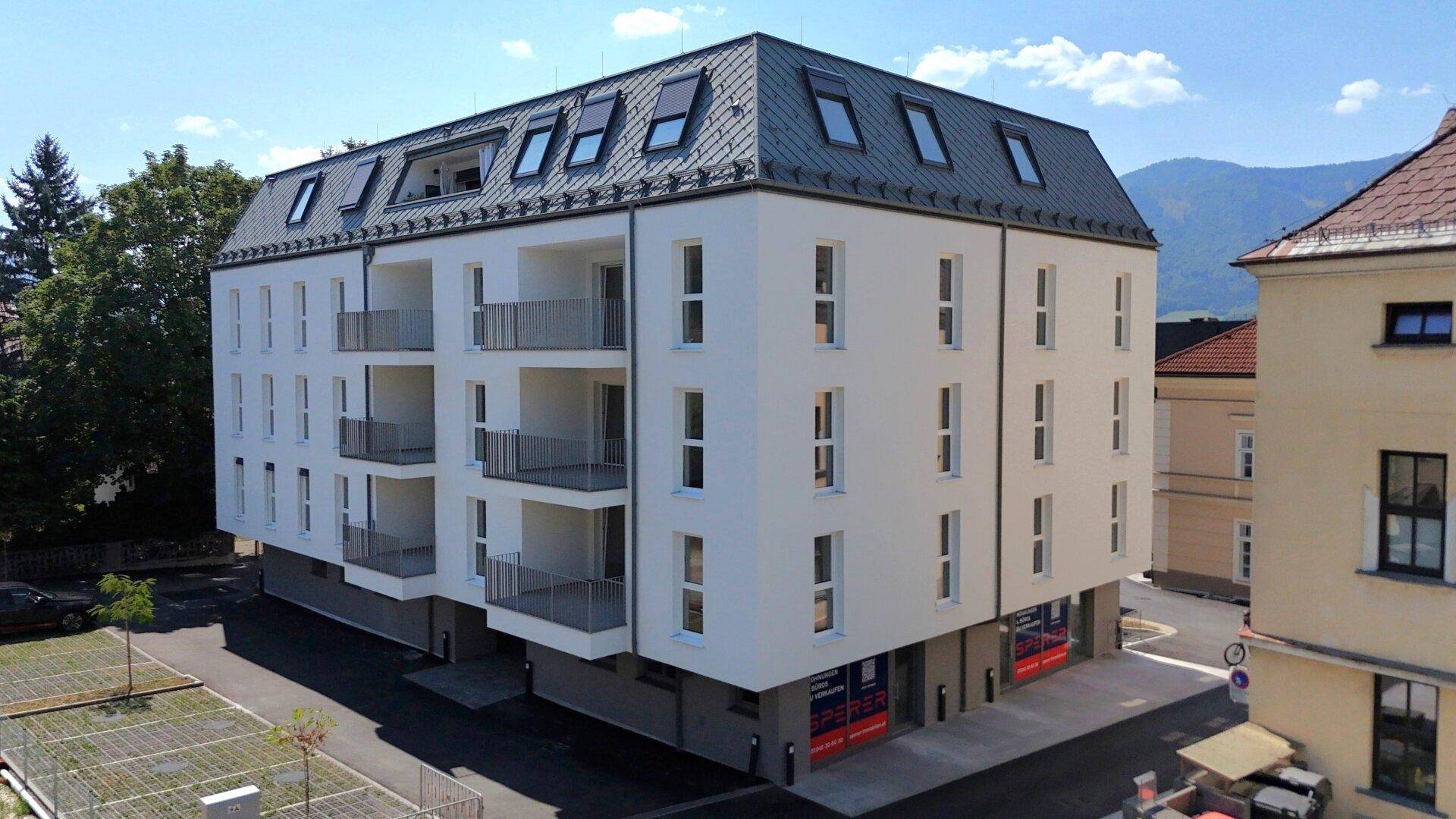 Modernes Wohngebäude mit weißer Fassade, Balkonen und einem markanten grauen Mansarddach.