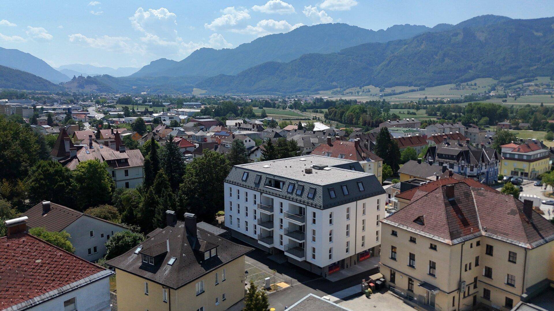 Luftaufnahme einer Stadt mit einem modernen Wohngebäude und malerischer Bergkulisse im Hintergrund.