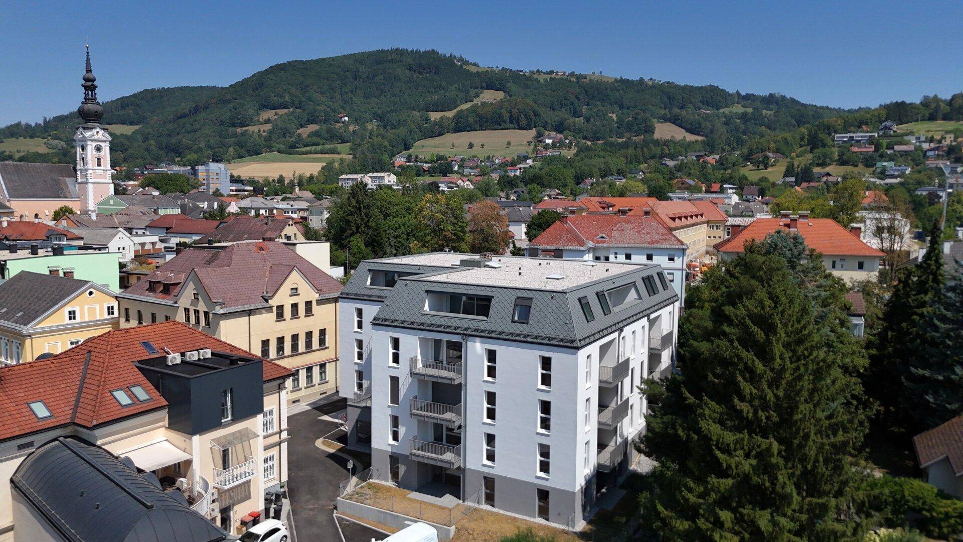 Panoramablick auf eine Stadt mit einem modernen Wohngebäude, umgeben von traditionellen Häusern und Hügeln.