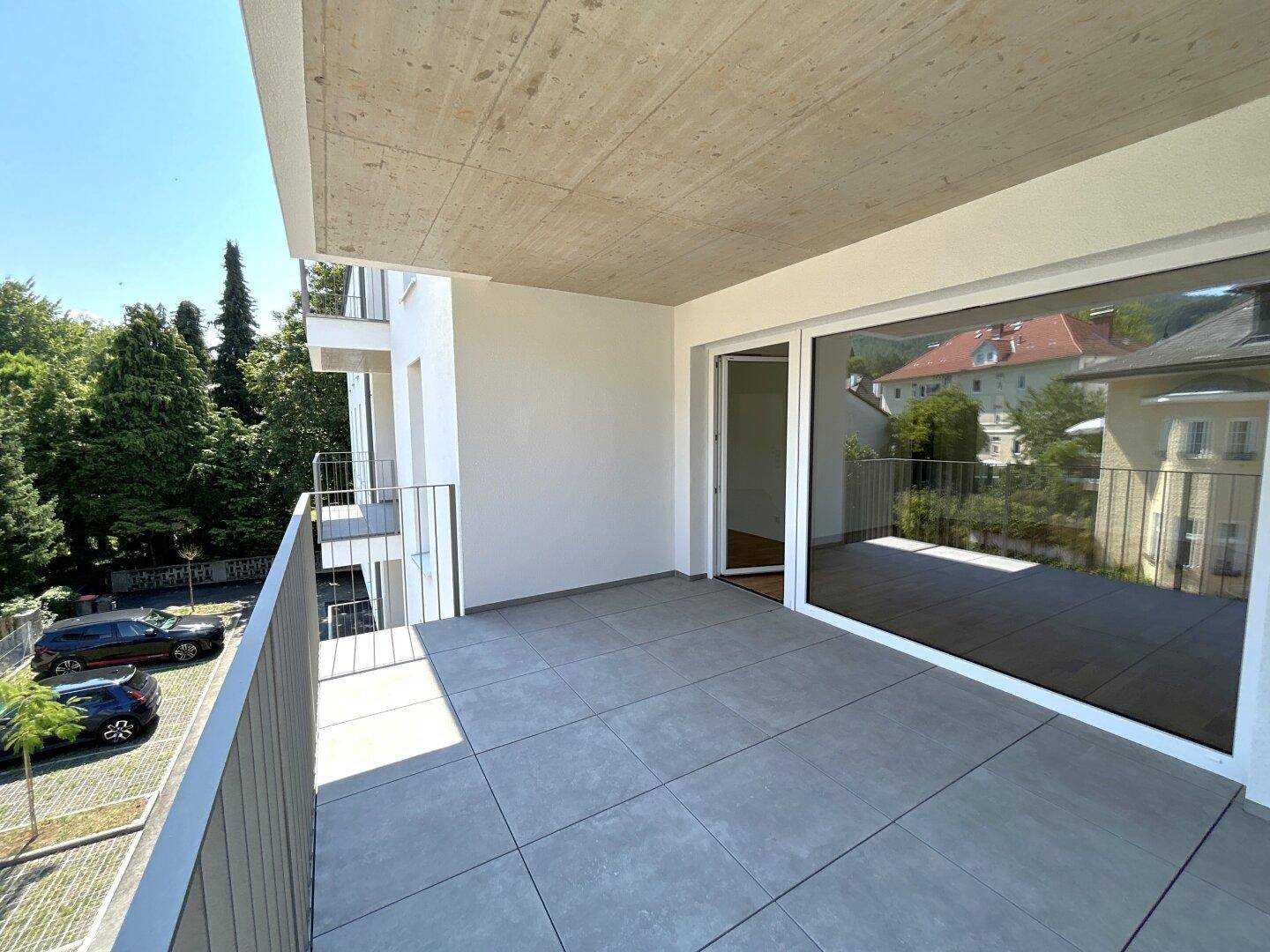 Moderner Balkon mit Fliesenboden und Sichtbetondecke, umgeben von grünen Bäumen und einem Parkplatz.