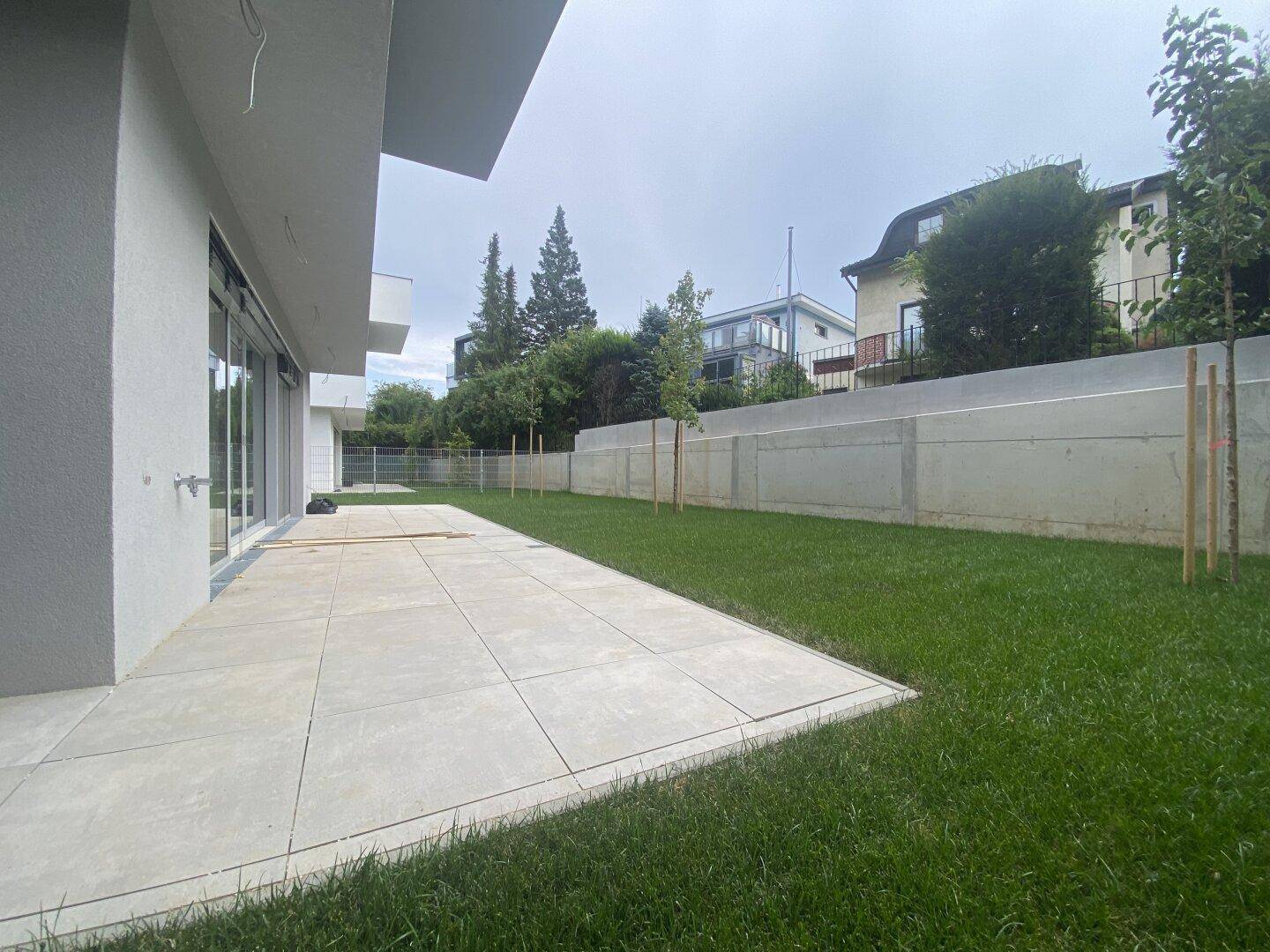 Großzügige, gepflasterte Terrasse mit angrenzendem Rasenbereich und einer Betonmauer im Hintergrund.