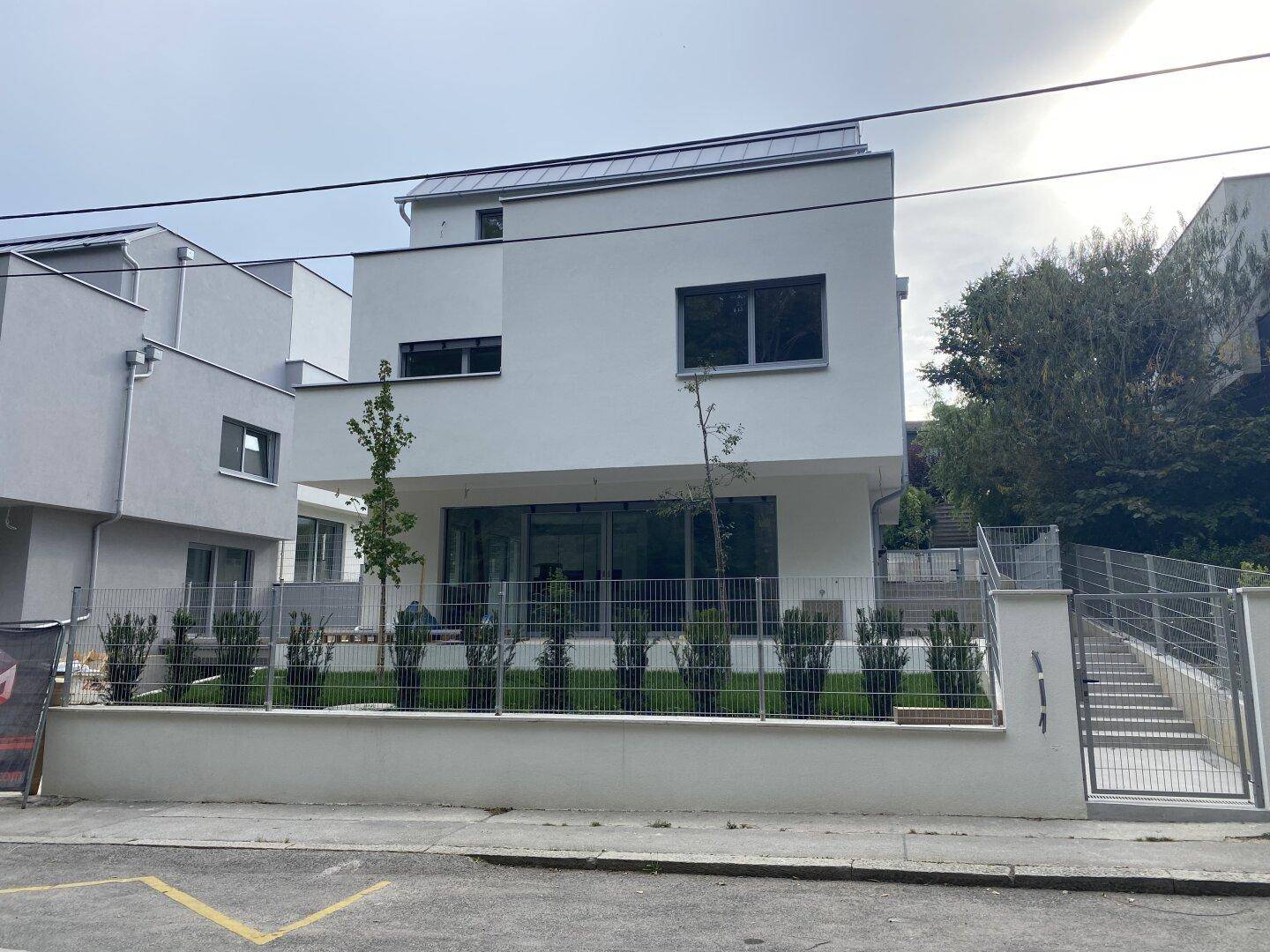 Moderne weiße Fassade eines Neubaus mit großen Fenstern und einem kleinen Vorgarten an der Straße.