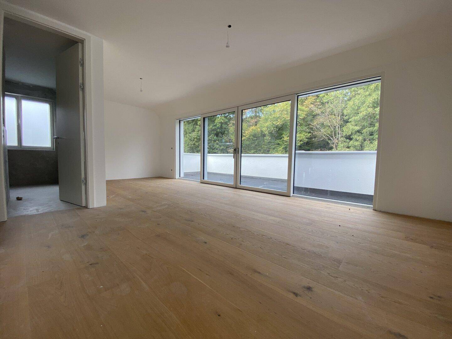 Geräumiger Innenraum mit Holzboden und Zugang zu einer Terrasse sowie einem weiteren unfertigen Raum.