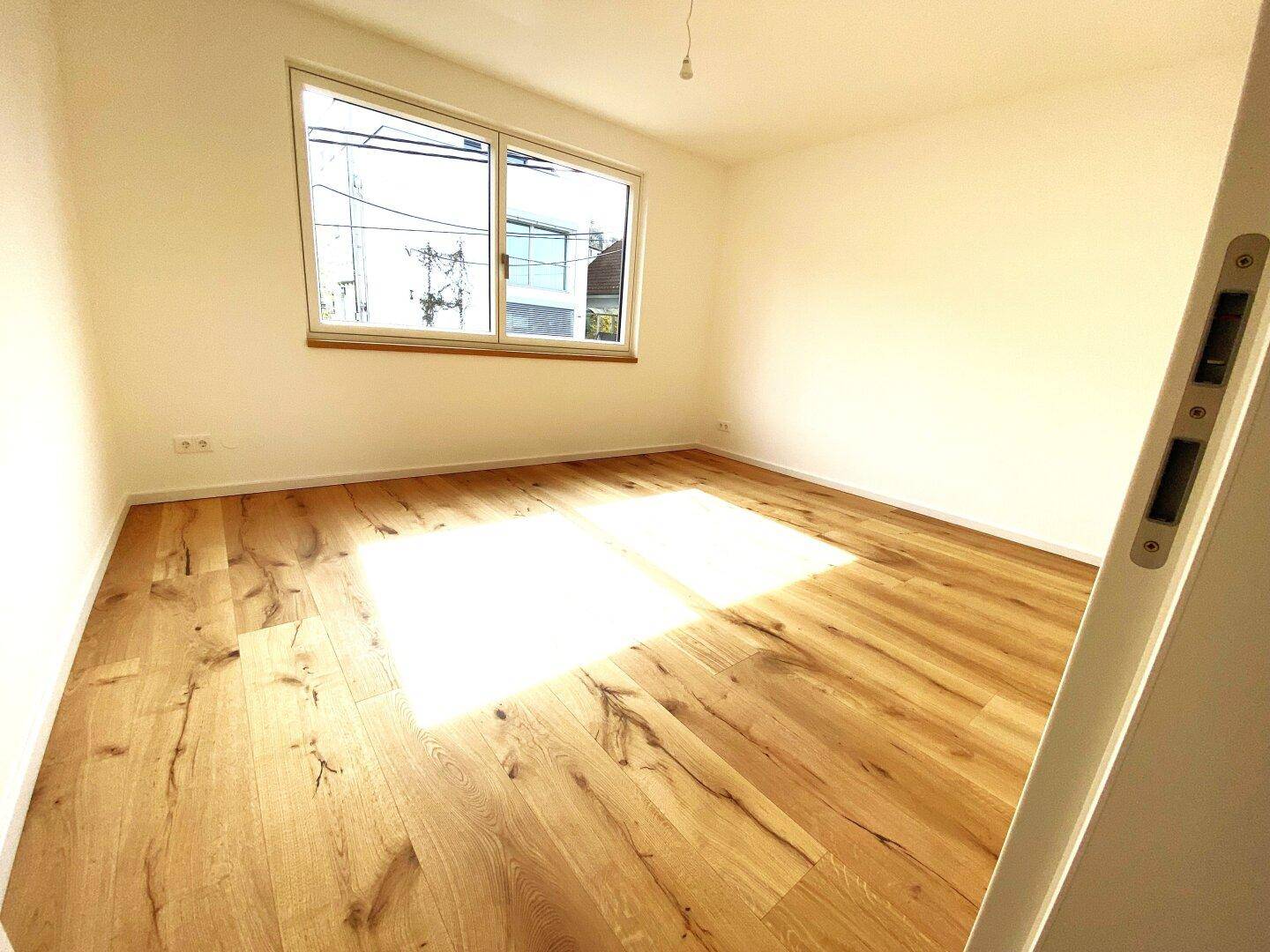 Sonniger Raum mit hochwertigem Holzparkett und einem großen Fenster, das viel Licht hereinlässt.