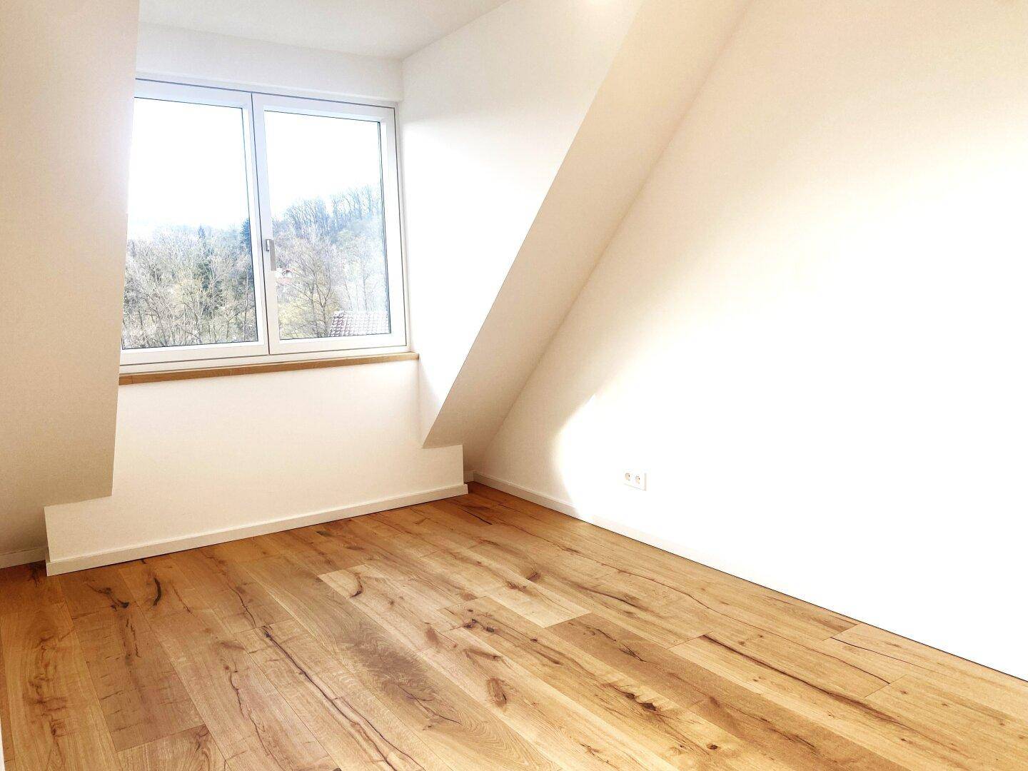 Gemütliches Zimmer im Dachgeschoss mit schrägen Wänden, Holzparkett und Fenster mit Ausblick.