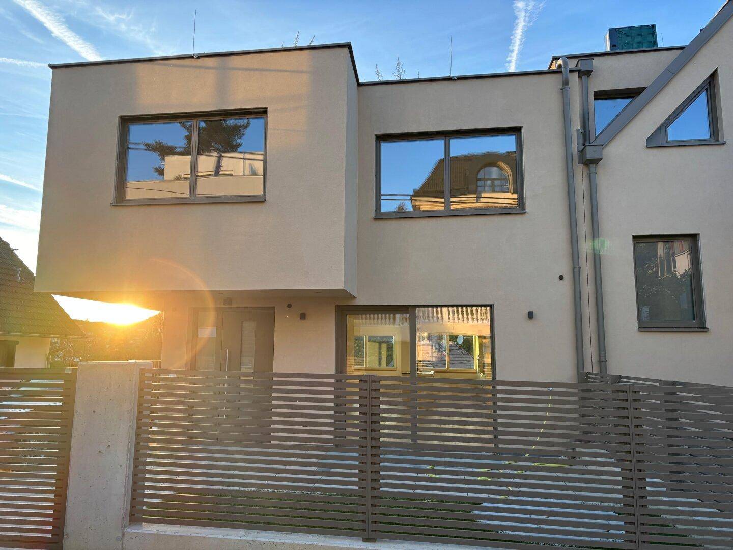 Moderne Gebäudefassade mit großen Fenstern und einem stilvollen Zaun im warmen Licht der Abendsonne.