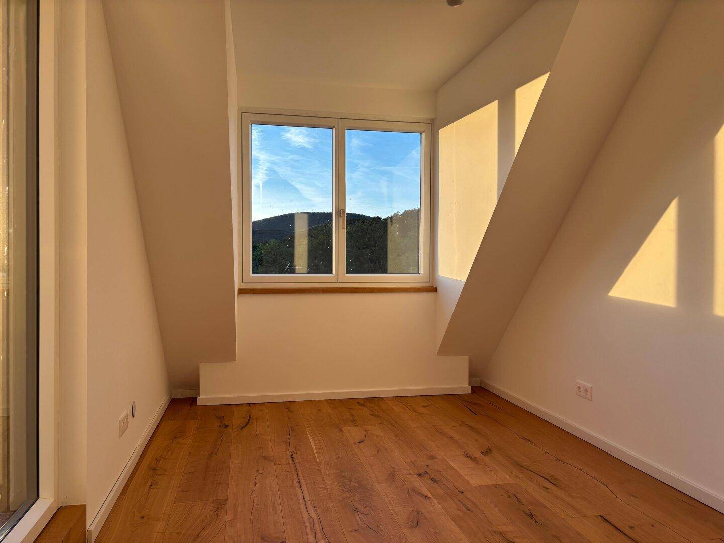 Helles Dachzimmer mit schrägen Wänden, Holzparkett und einem Fenster mit Blick ins Grüne.