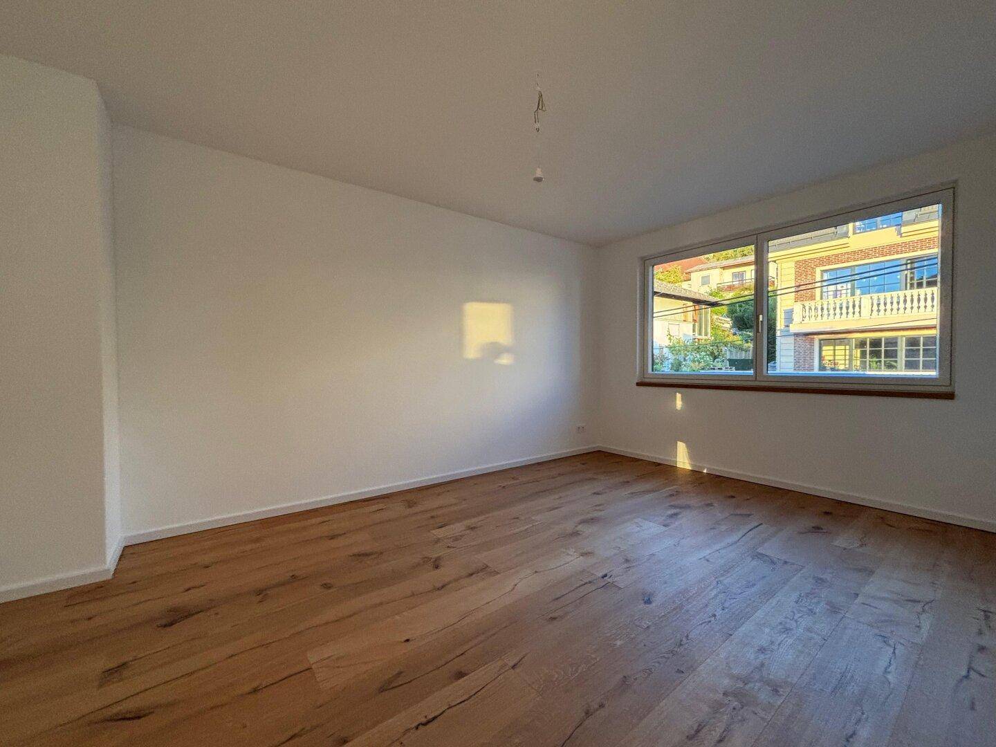 Helles Zimmer mit Holzparkett und einem großen Fenster, das den Raum mit Sonnenlicht füllt.
