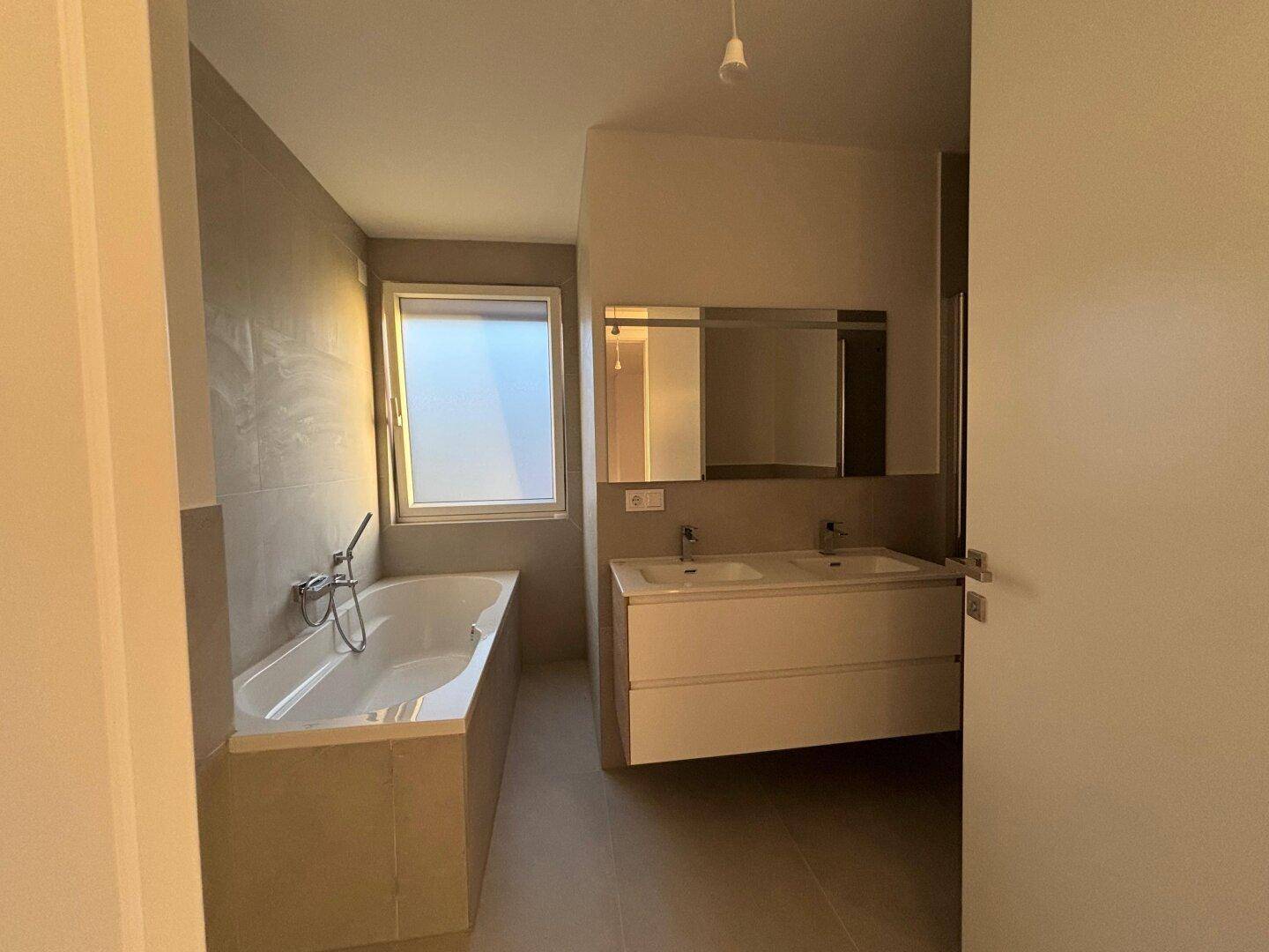 Modernes Badezimmer mit Badewanne, Doppelwaschbecken und Fenster, hell gefliest.