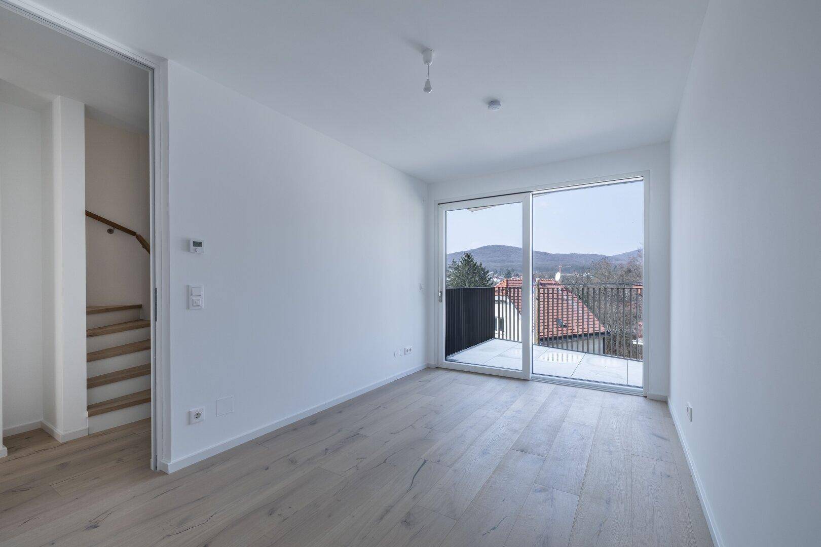 Helles Zimmer mit Holzboden, Balkonzugang und weitem Blick über die Dächer und Landschaft.