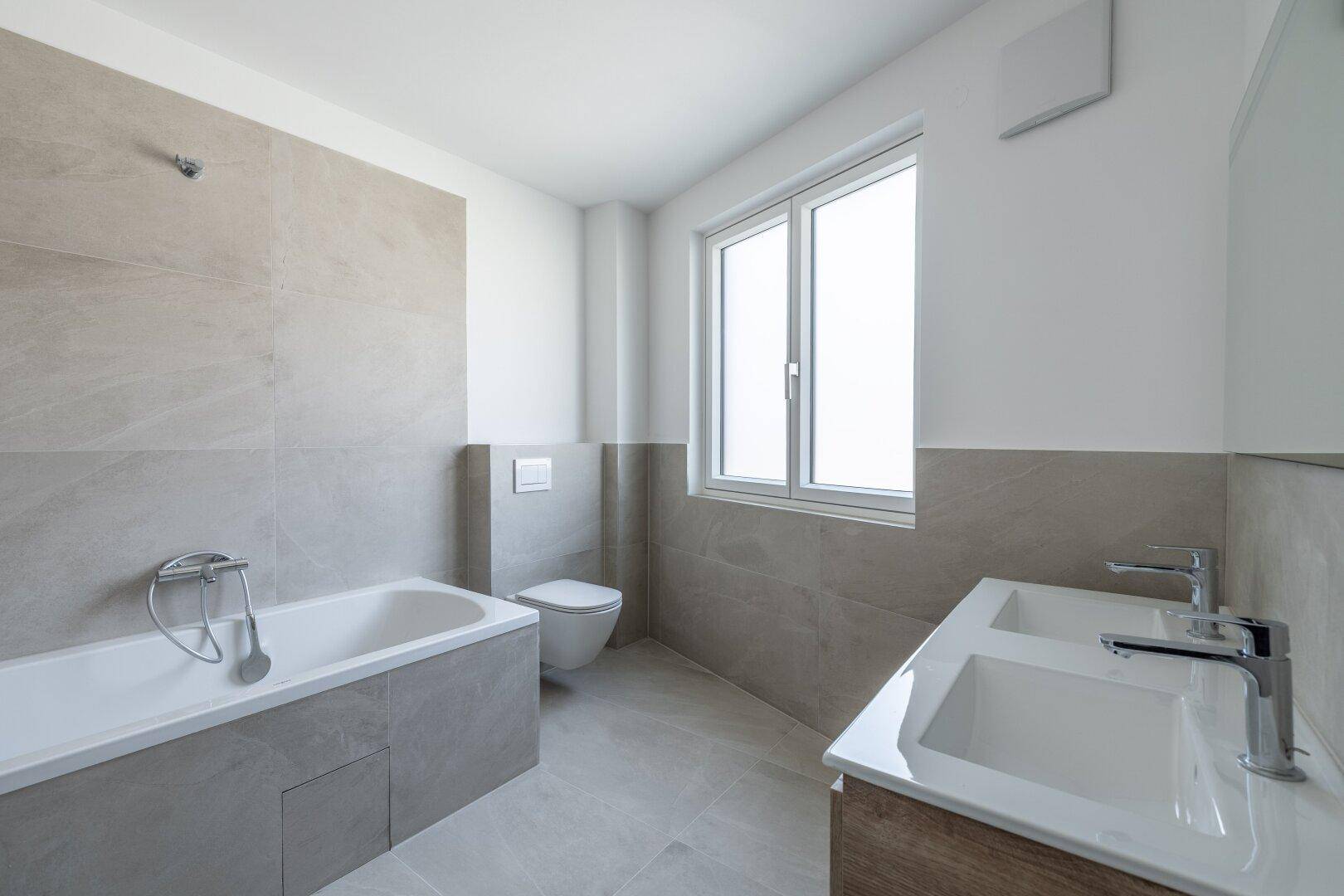 Modernes Badezimmer mit Badewanne, Doppelwaschbecken und Wand-WC in hellen Grautönen.