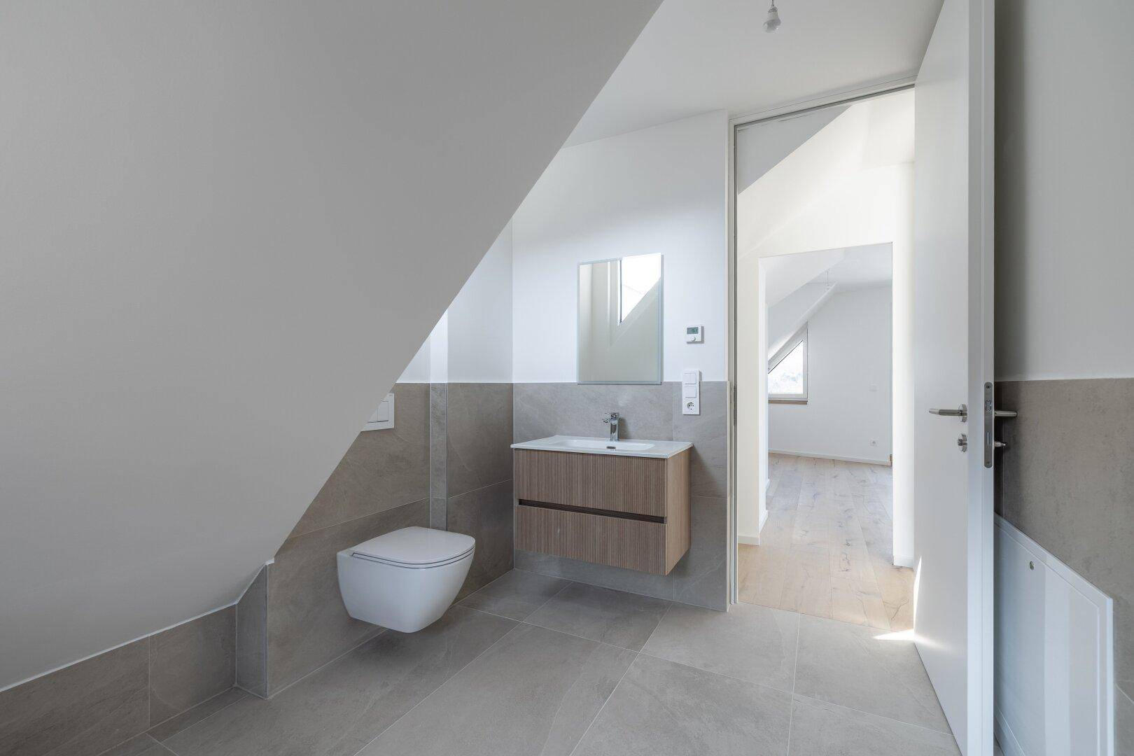 Helles Dachgeschoss-Badezimmer mit Waschtisch, Wand-WC und Spiegel unter einer Dachschräge.