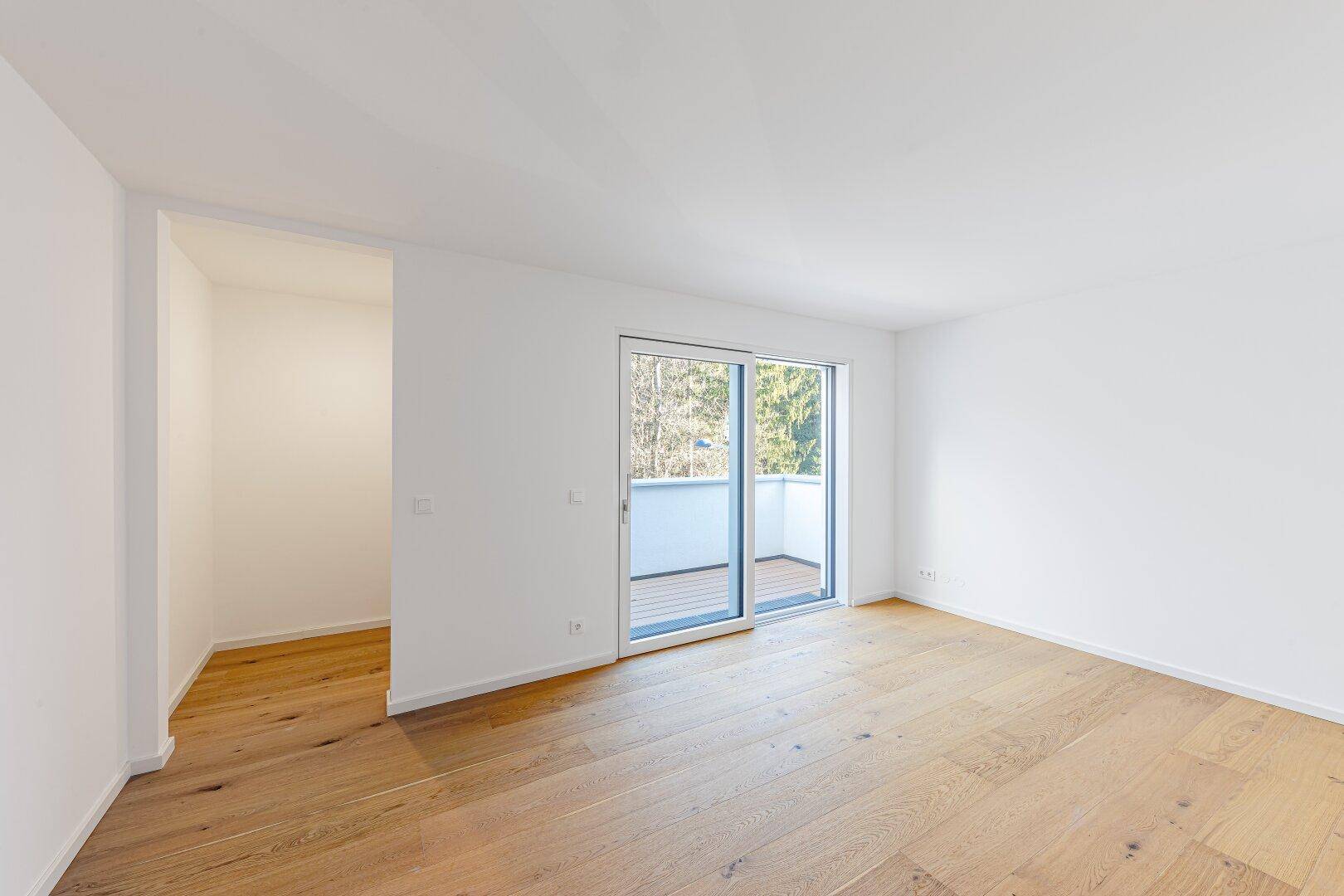Helles Zimmer mit Holzboden und Schiebetür, die zu einem Balkon mit Blick ins Grüne führt.