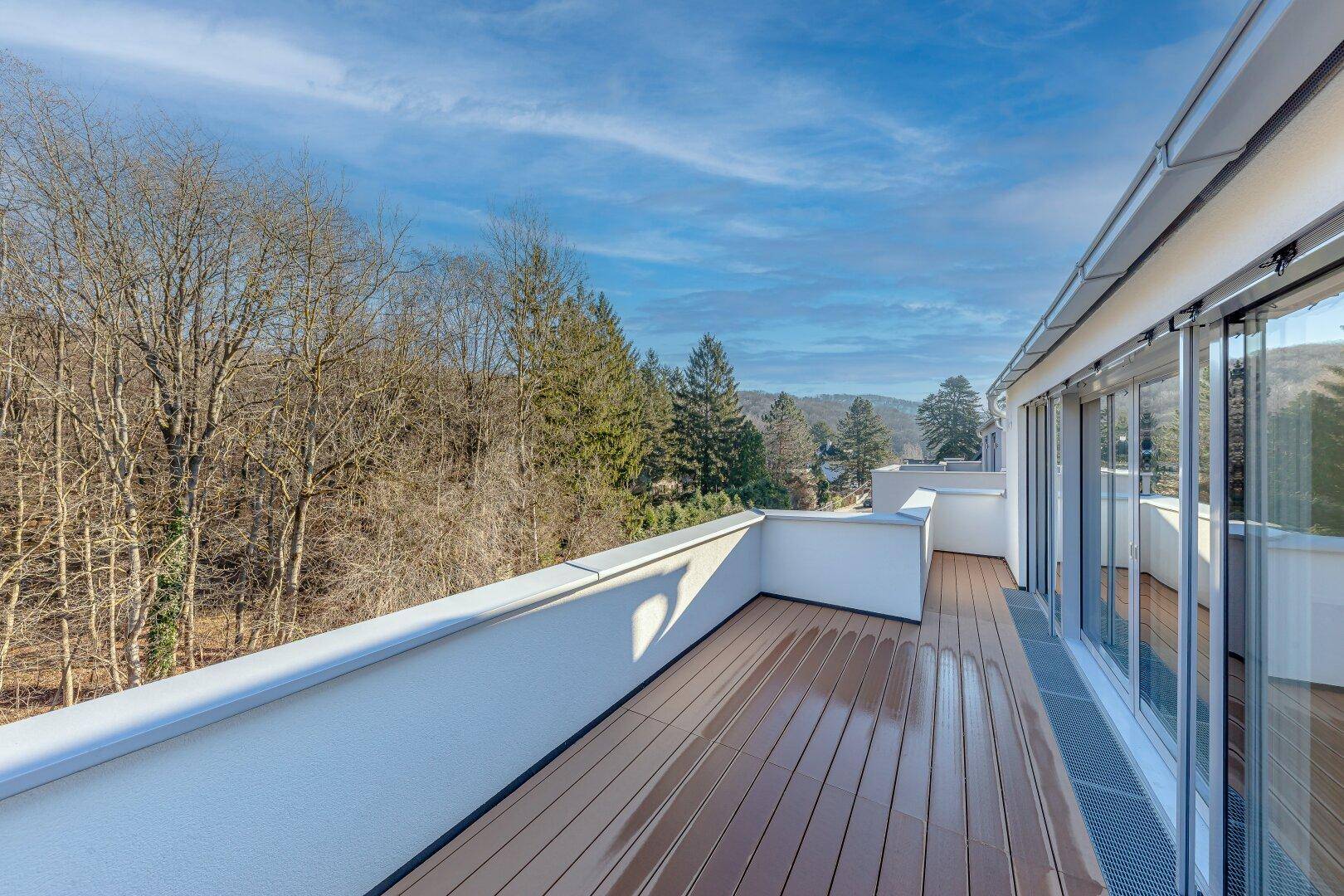 Geräumige Terrasse mit modernem Holzboden und weitem Blick auf die umliegende Natur und bewaldete Hügel unter blauem Himmel.