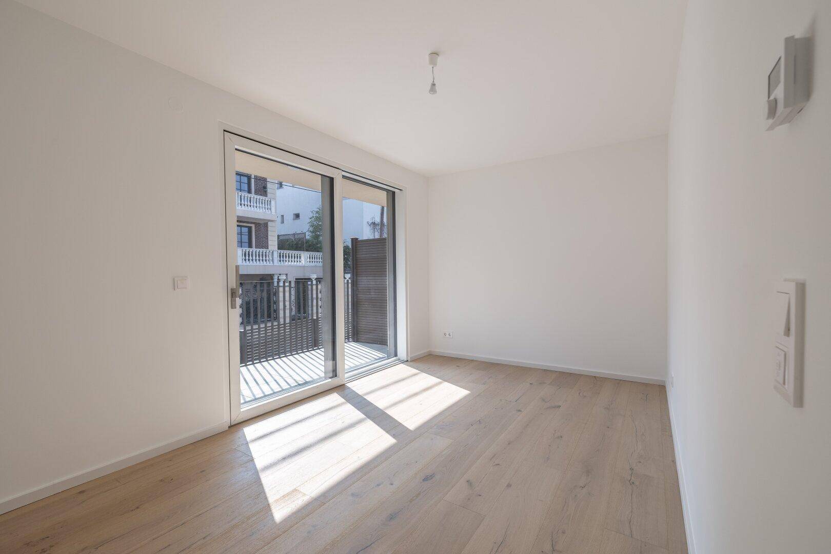 Helles Zimmer mit Holzboden und großem Schiebetürzugang zu einem Balkon, durchflutet von Sonnenlicht.