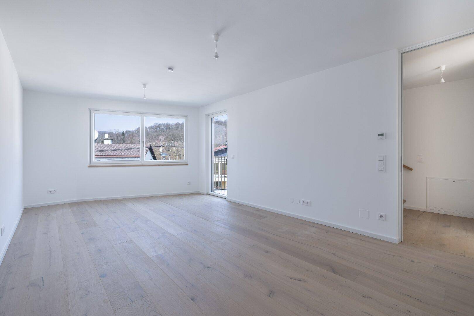 Großzügiger, heller Wohnraum mit Holzboden und Fenstern, die viel Licht hereinlassen und einen Ausblick bieten.
