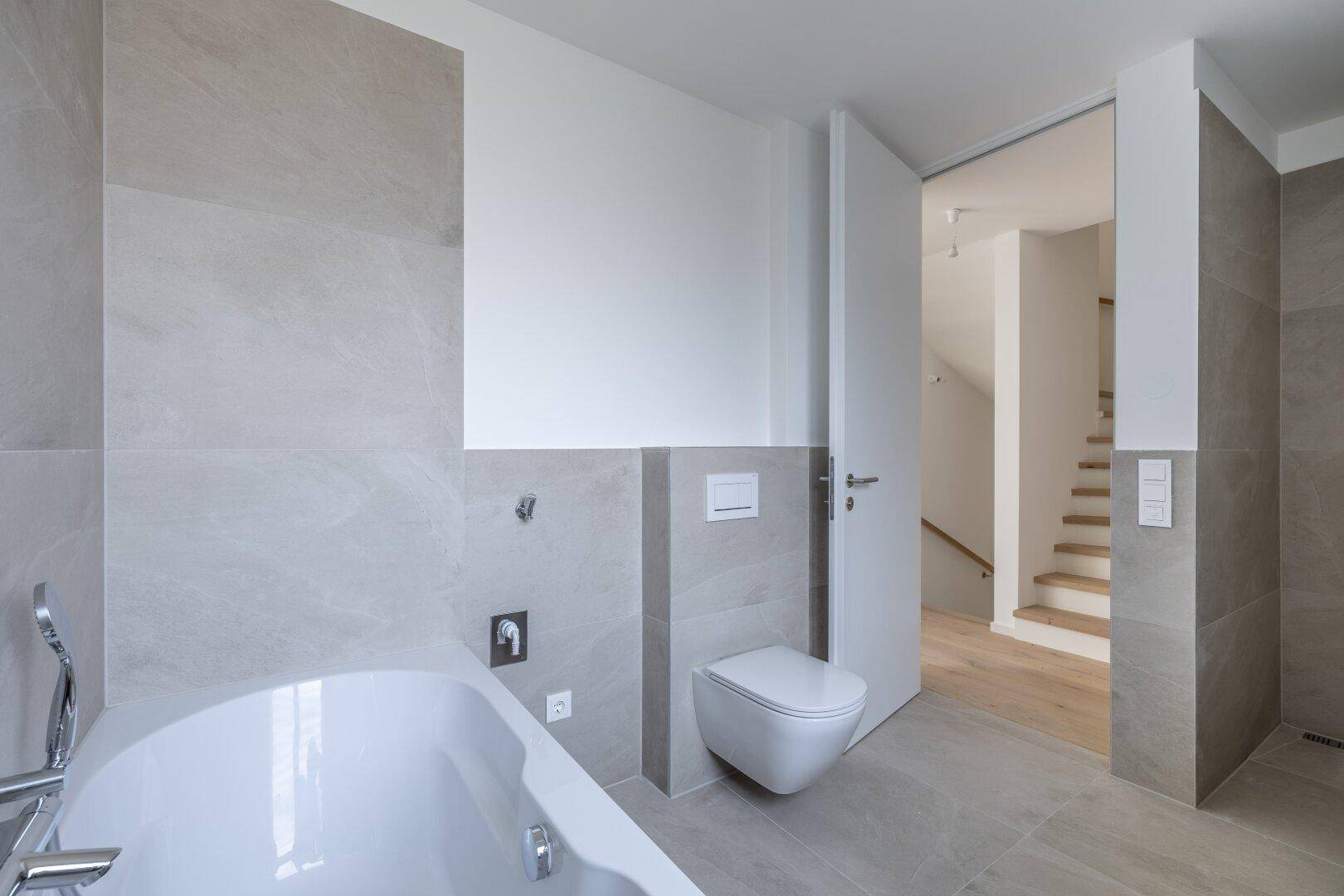 Modernes Badezimmer mit hellgrauen Fliesen, wandhängendem WC und integrierter Badewanne, sehr gepflegt.