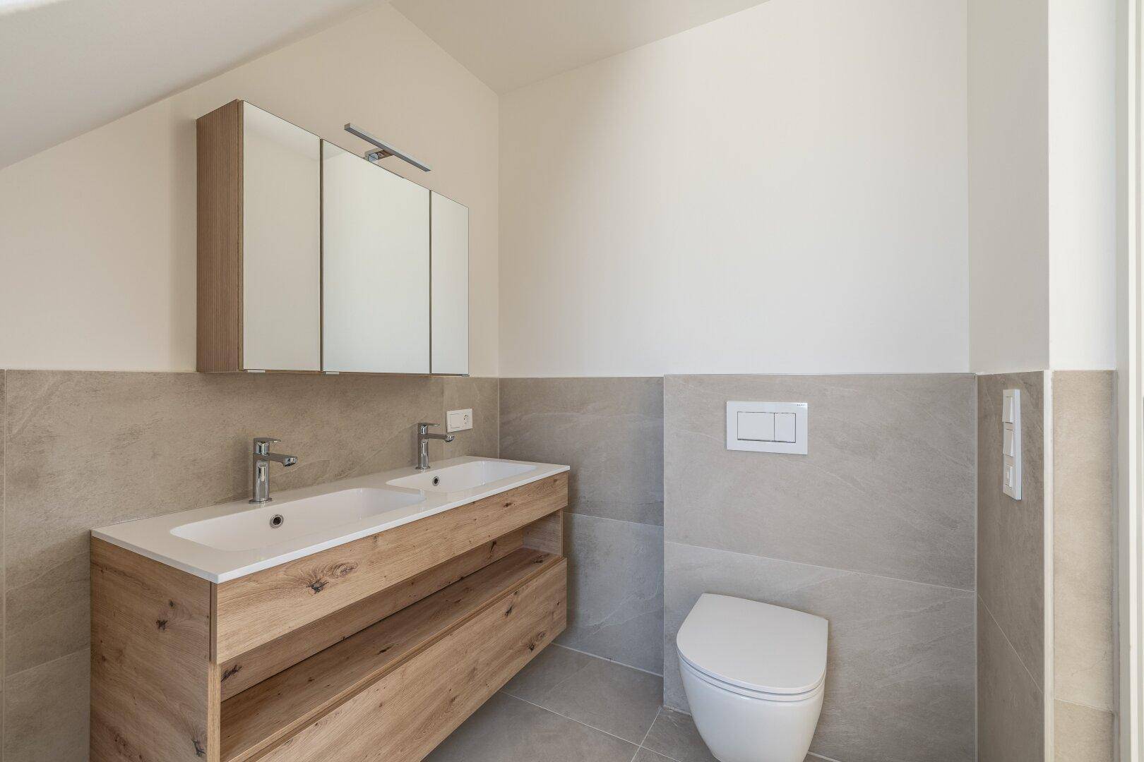 Modernes Badezimmer mit Doppelwaschbecken, Holzunterschrank und wandhängendem WC unter einer Dachschräge.