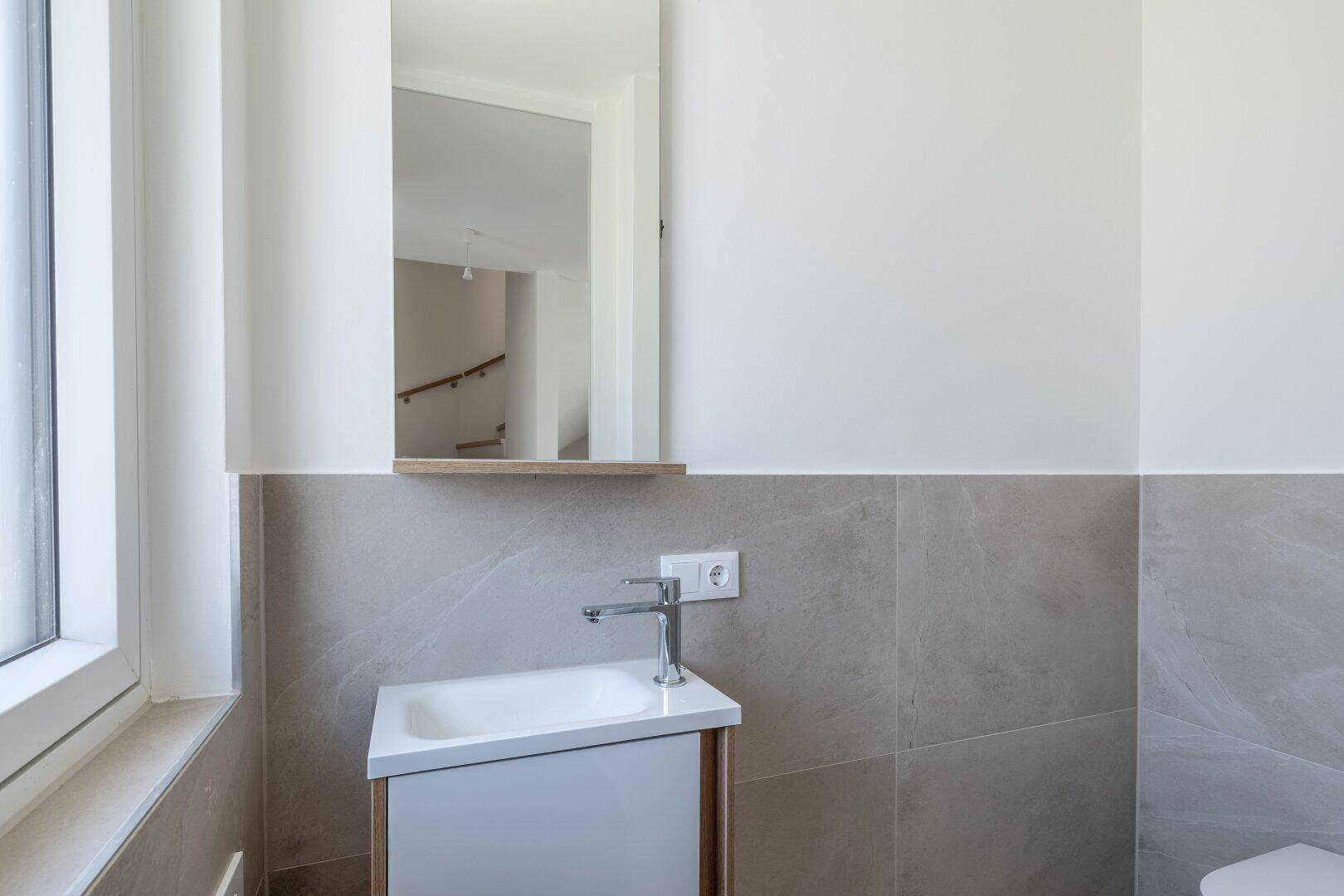 Kleines Badezimmer mit Einzelwaschbecken, Spiegel und Fenster, modern gefliest und funktional.