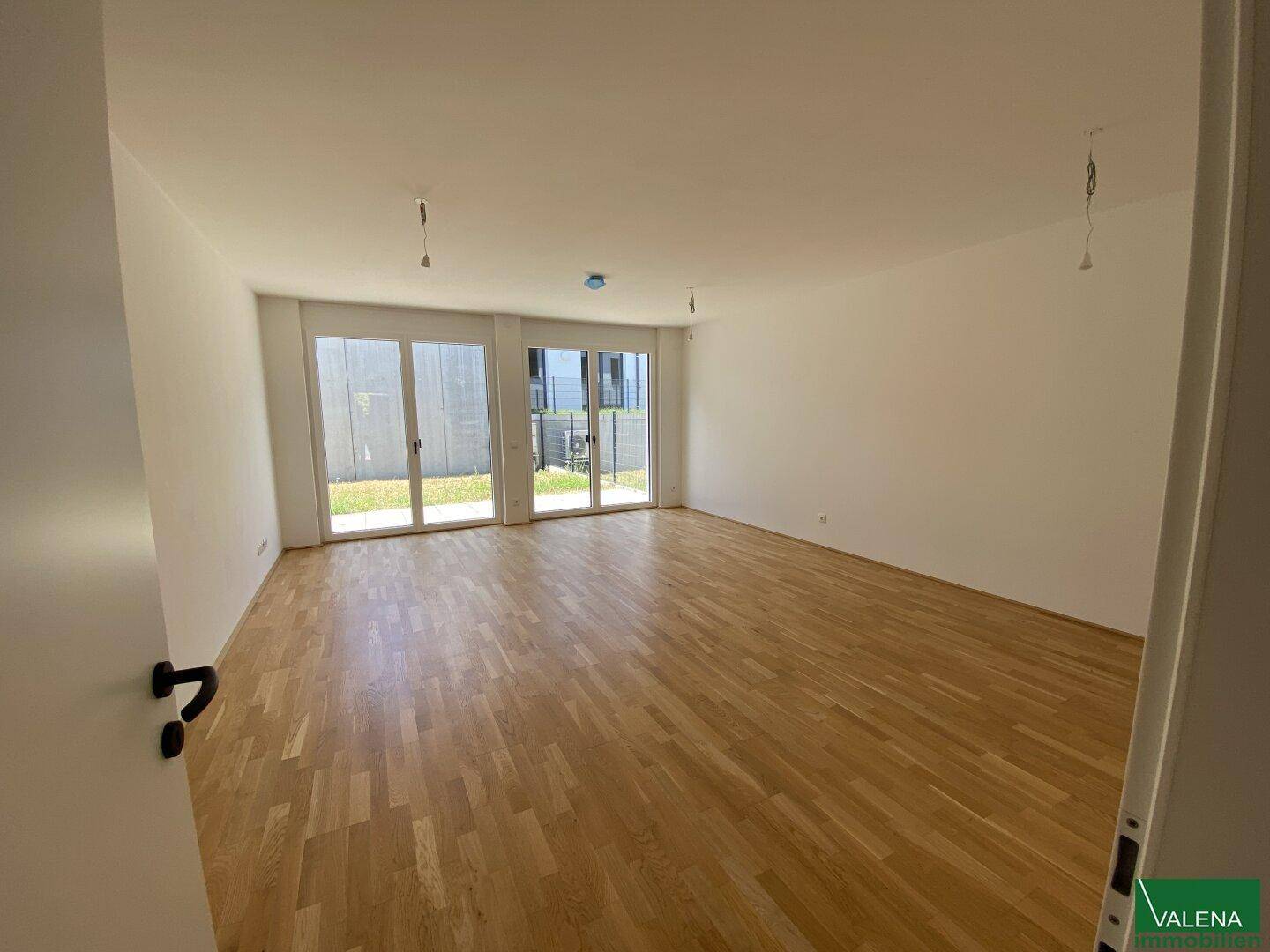 Helles, leeres Wohnzimmer mit Holzboden und großen Fenstern zum Gartenbereich.