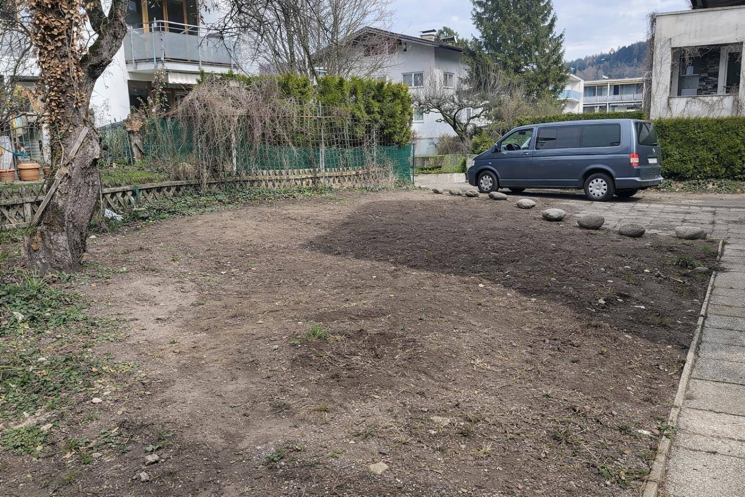 Vorbereiteter Gartenbereich mit einem geparkten Fahrzeug und umgebenden Bäumen sowie Hecken.