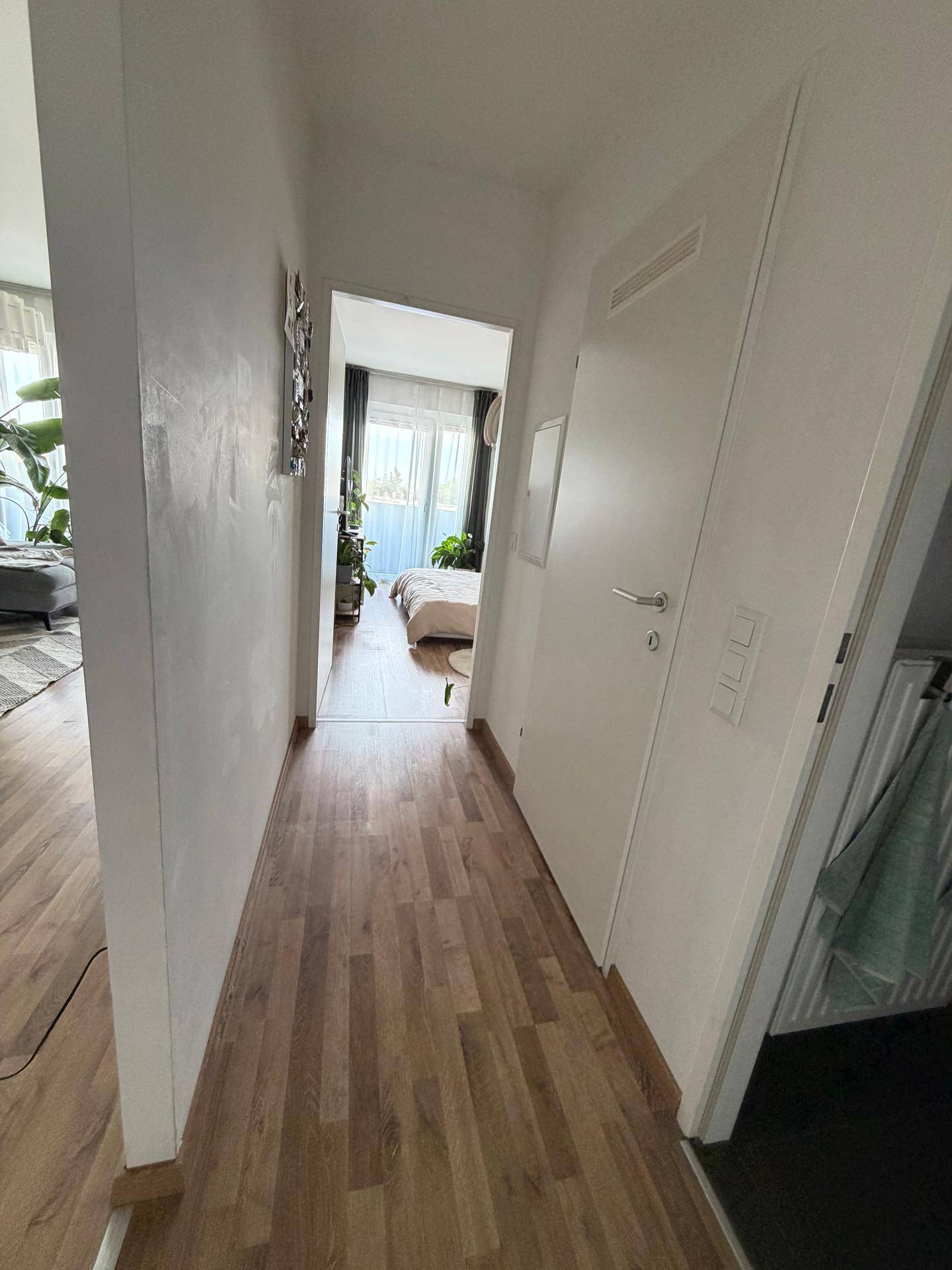 Flur mit Holzboden und Blick in ein helles Schlafzimmer mit Balkonzugang.