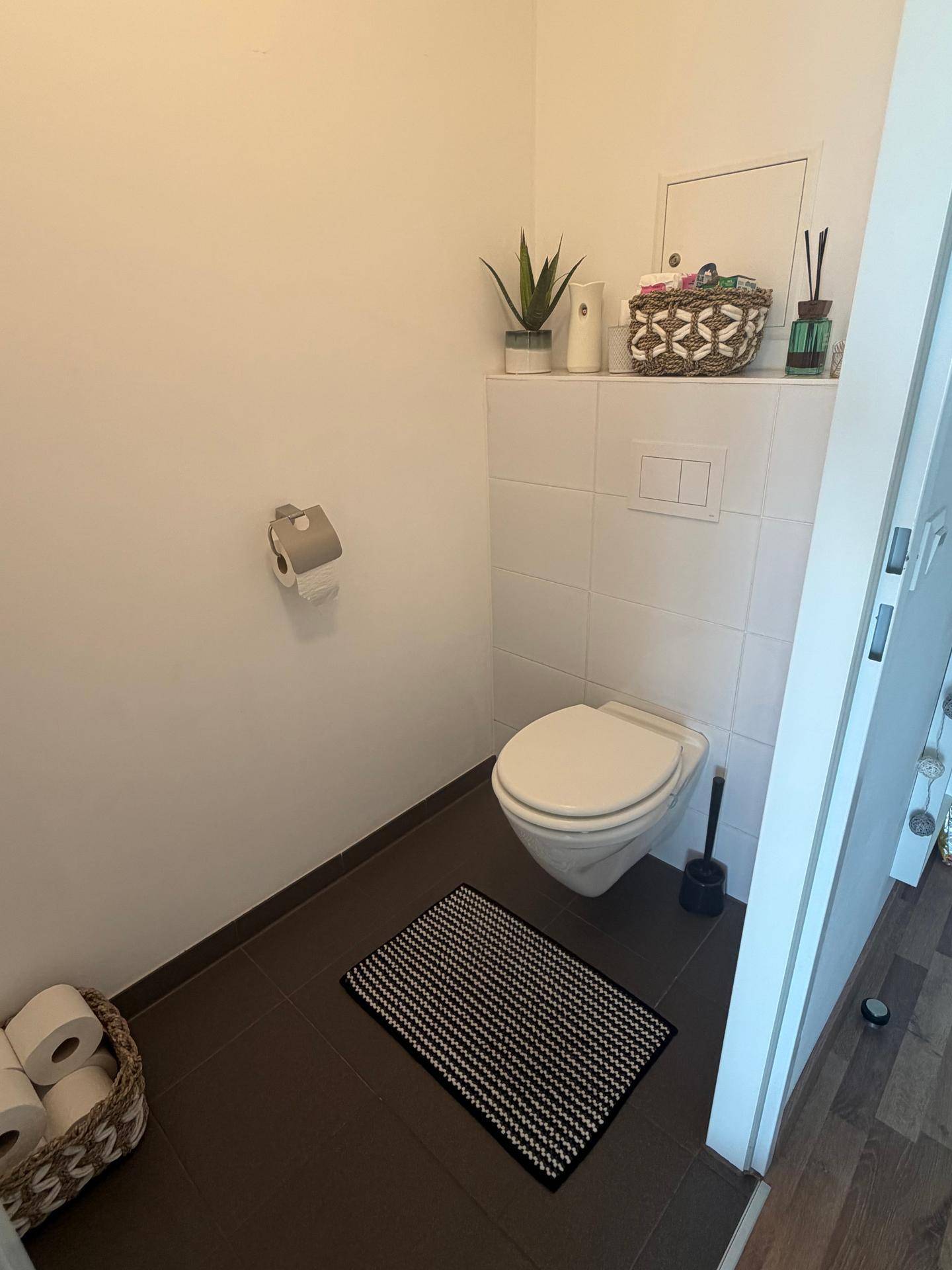 Separates WC mit wandhängender Toilette und dunklem Fliesenboden.