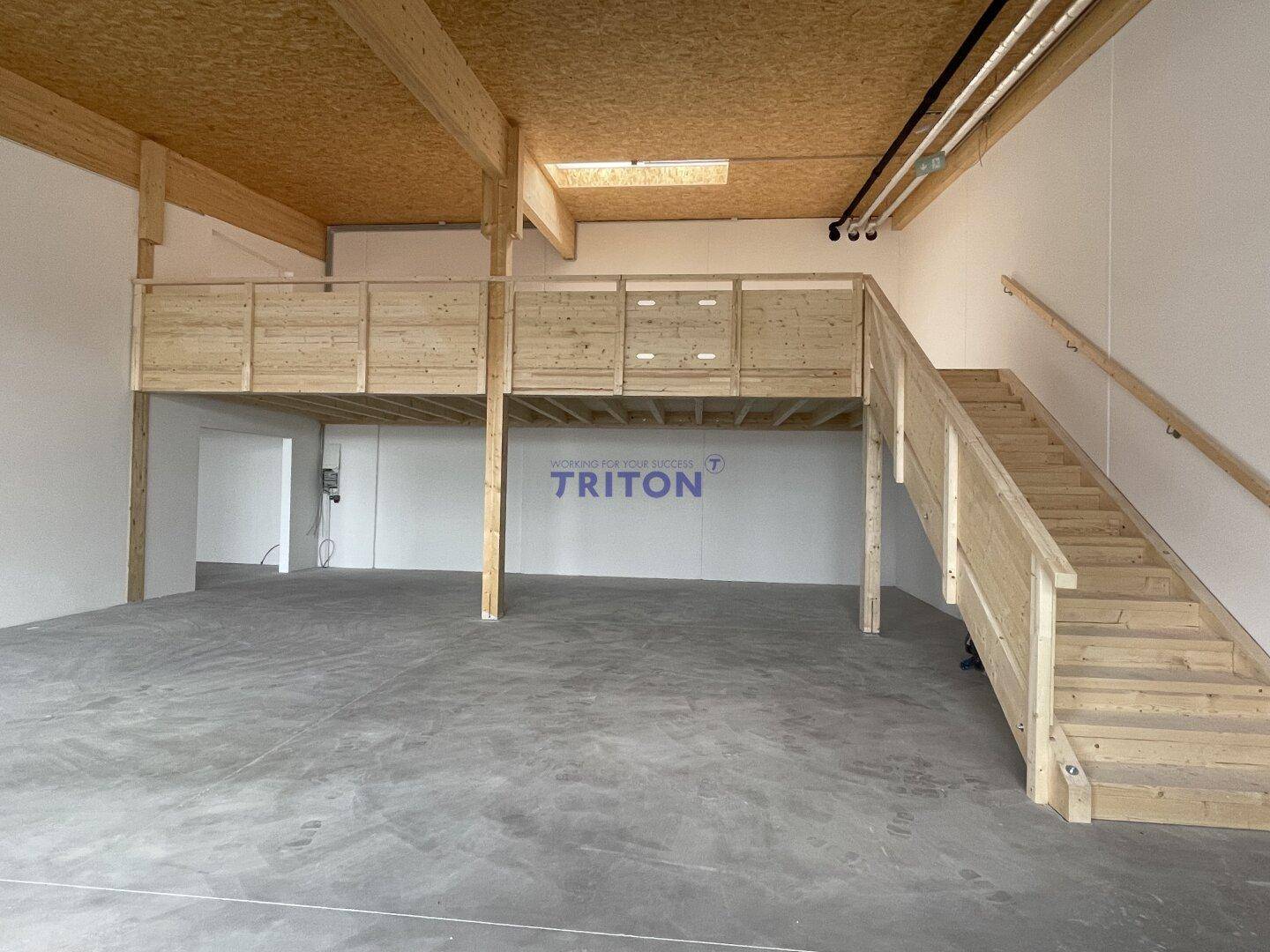 Große Gewerbefläche mit Betonboden, Holzgalerie und Holztreppe, viel Tageslicht durch Dachfenster.