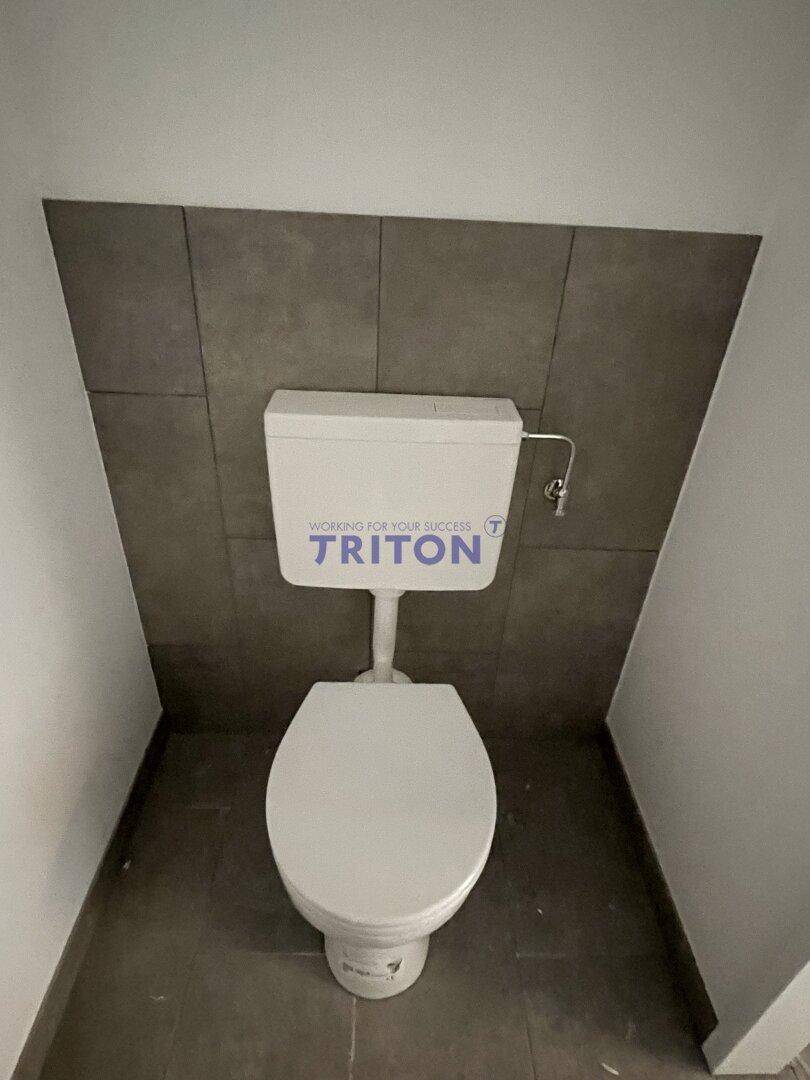 Einfache Toilette mit Fliesen an der Wand und auf dem Boden in neutralen Grautönen.