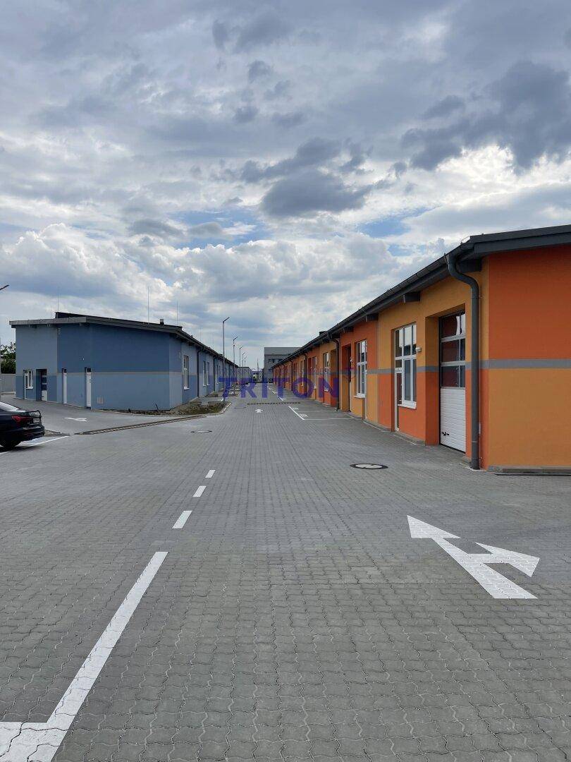 Außenansicht einer Reihe von Gewerbegebäuden mit gepflasterter Zufahrt und Parkplätzen unter bewölktem Himmel.