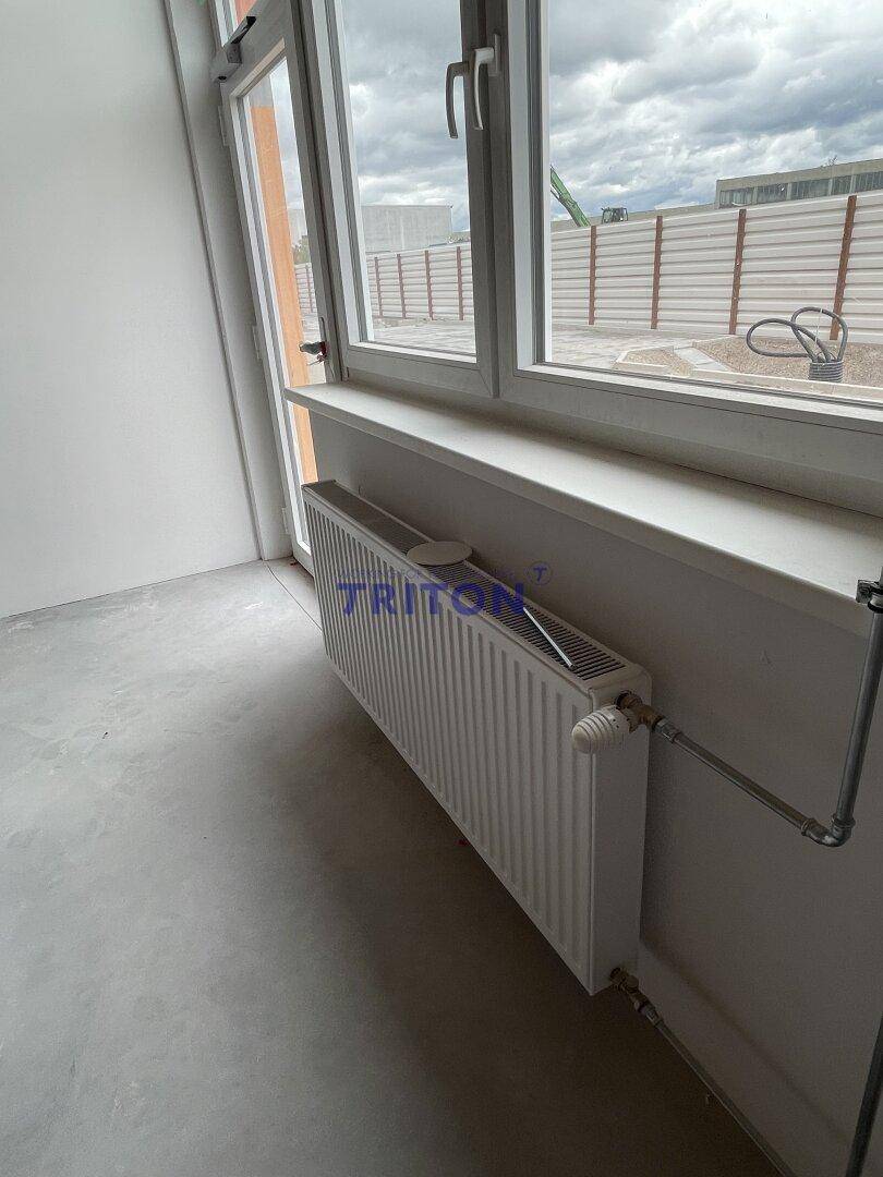 Heizkörper unter einem Fenster in einem hellen Raum mit Betonboden und weißer Wand.
