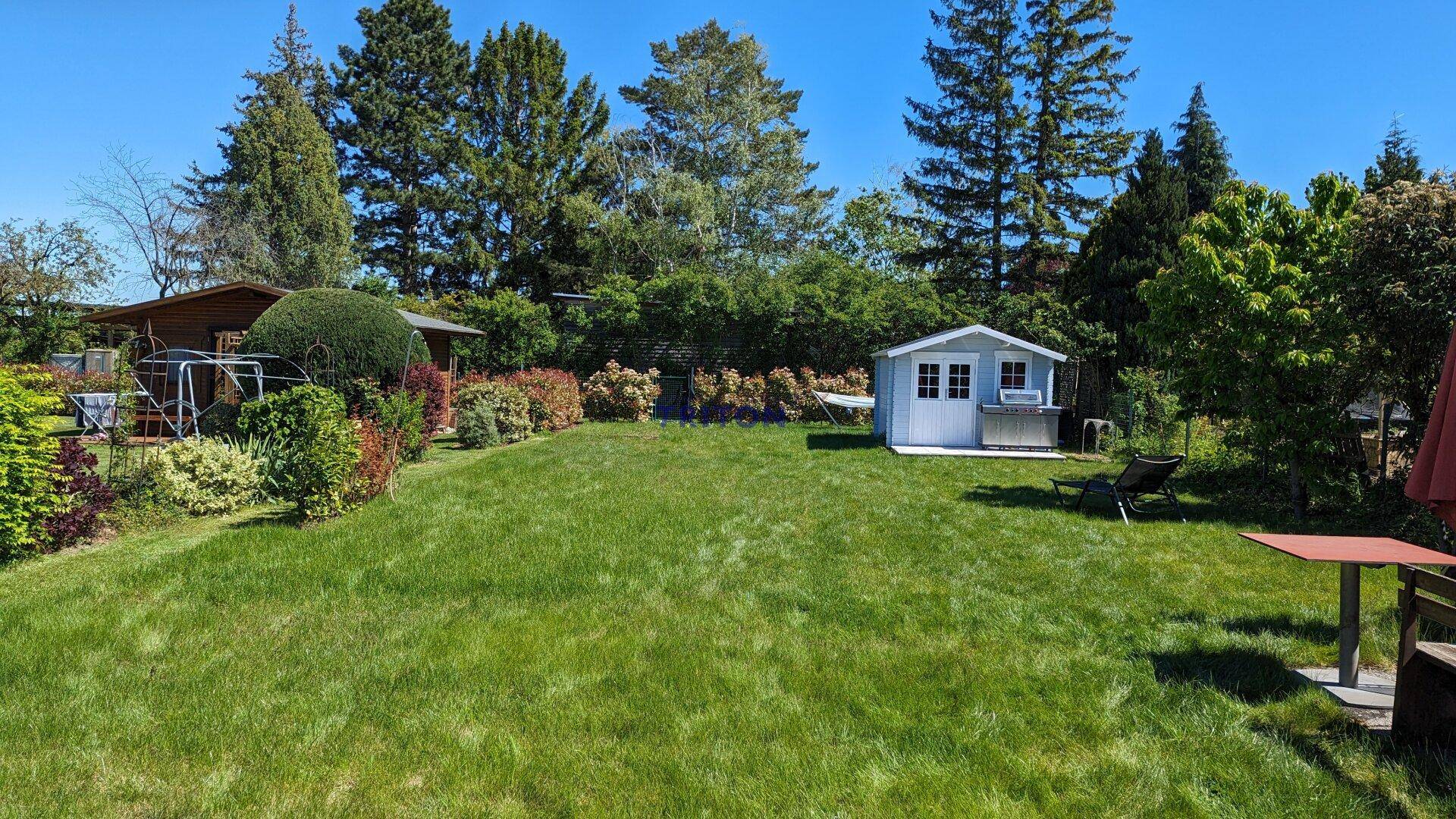 Großer grüner Garten mit Rasenfläche, einem Gartenhaus und Grillbereich unter blauem Himmel.
