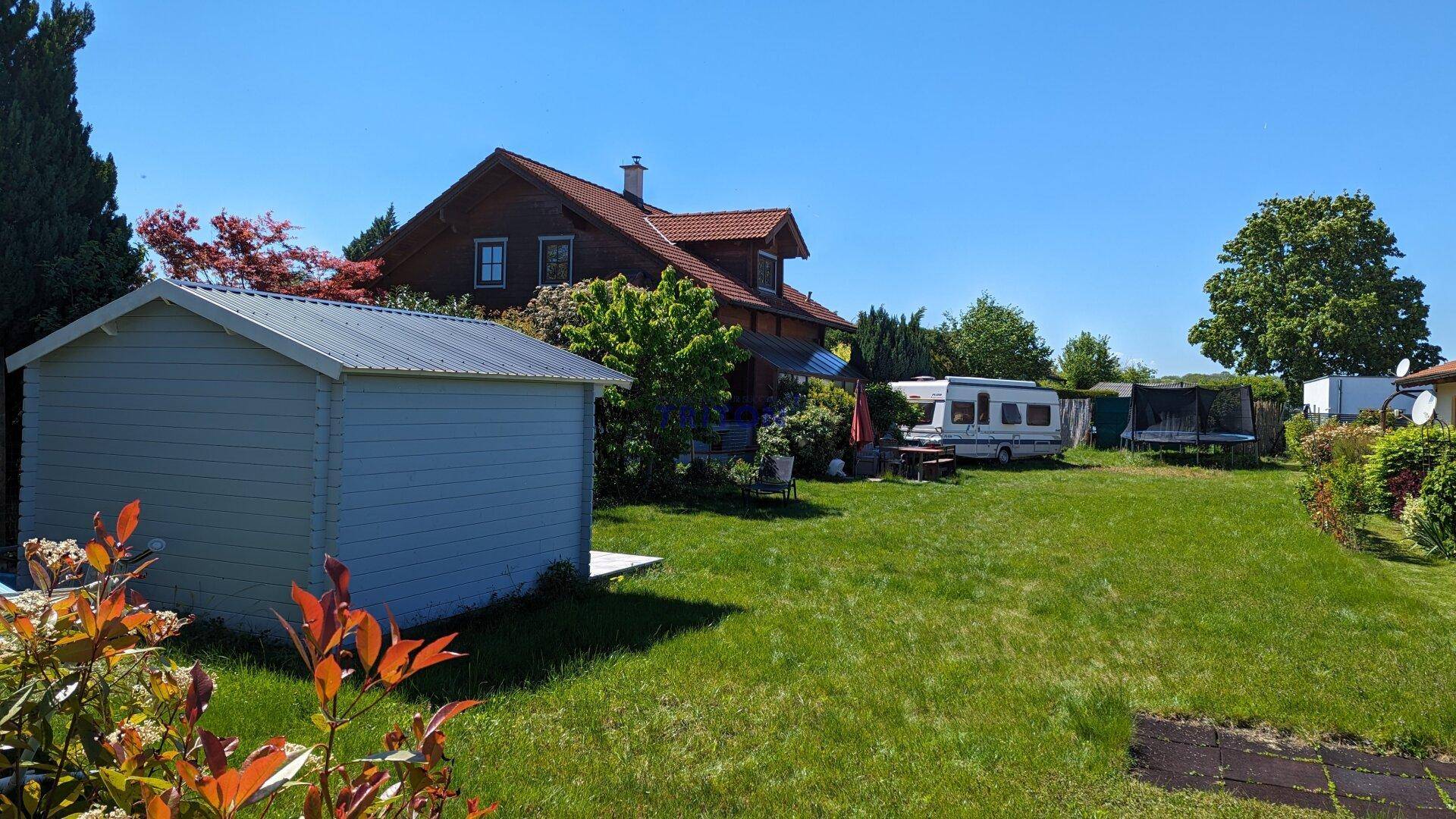 Großer Garten mit Wohnhaus, Wohnwagen, Trampolin und Gartenhaus, umgeben von viel Grün.