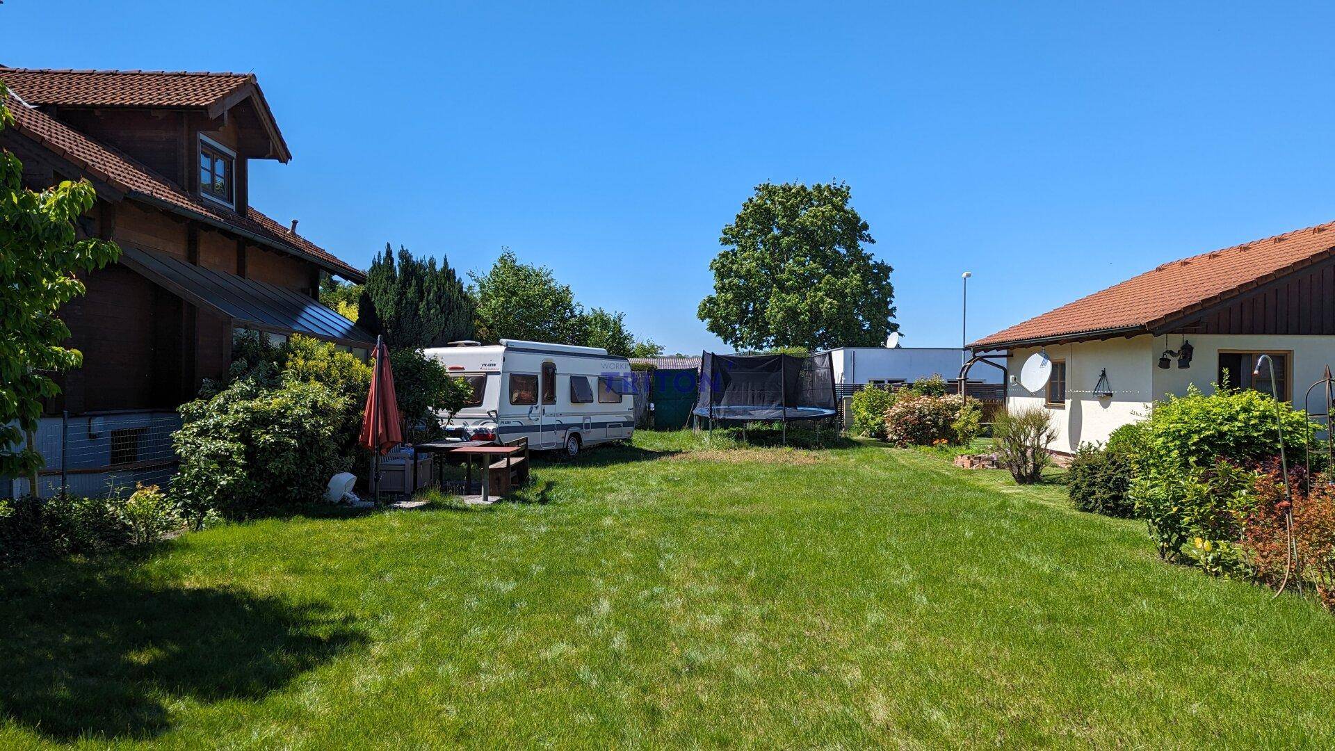 Weitläufiger Gartenbereich mit zwei Häusern, einem Wohnwagen und einem Trampolin unter blauem Himmel.