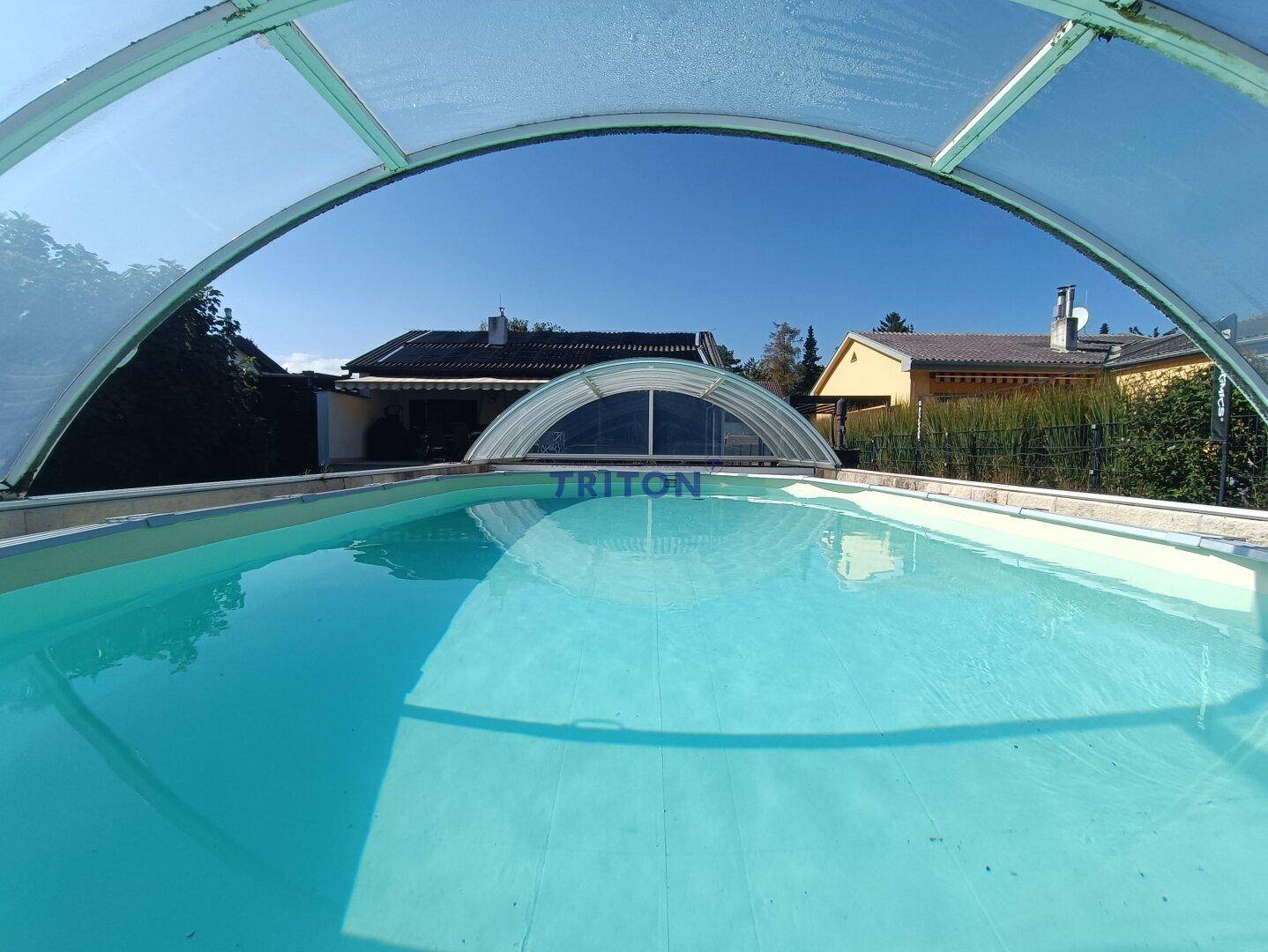 Überdachter Swimmingpool mit klarem Wasser und Blick auf die umliegenden Häuser und Bäume.