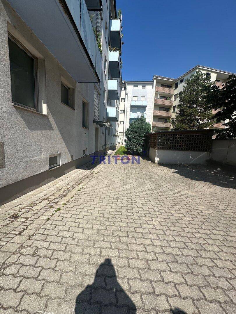 Gepflasterter Parkplatzbereich neben dem Gebäude mit weiteren Wohnhäusern im Hintergrund.
