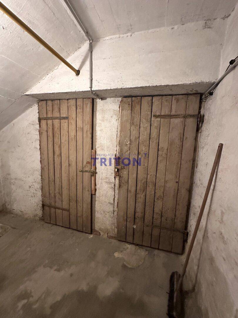 Kellerraum mit rauen Betonwänden, Holztüren und sichtbaren Rohren an der Decke.