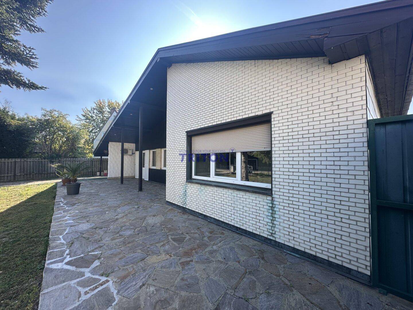 Seitlicher Blick auf das Haus mit gepflasterter Terrasse, weißer Ziegelfassade und Rollläden.