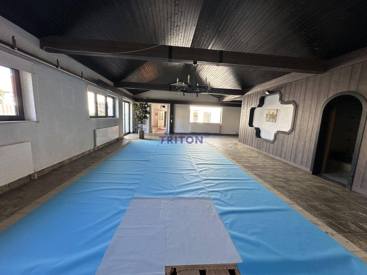 Weitläufiger Innenraum mit überdachtem Pool, Fliesenboden und Holzbalken an der Decke.