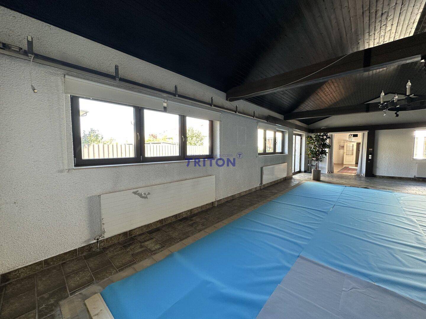 Großer Innenraum mit abgedecktem Pool, dunkler Holzdecke und Fenstern mit Blick ins Grüne.