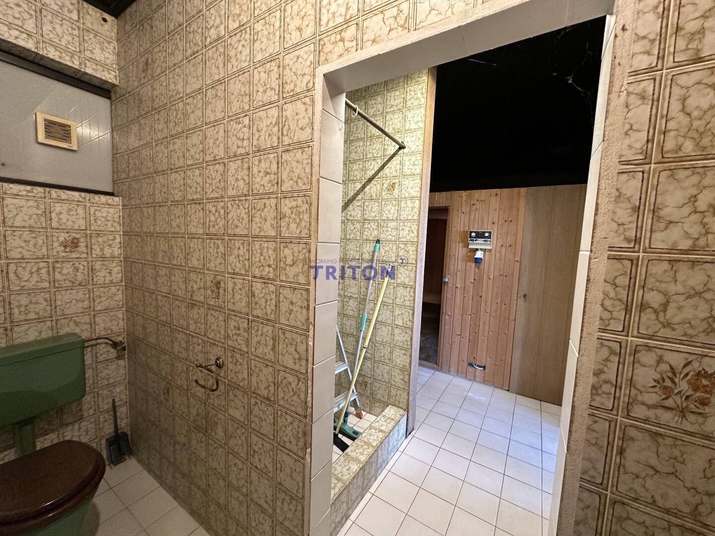 Badezimmer mit gemusterten Fliesen, grüner Toilette und Zugang zum Sauna- und Duschbereich.