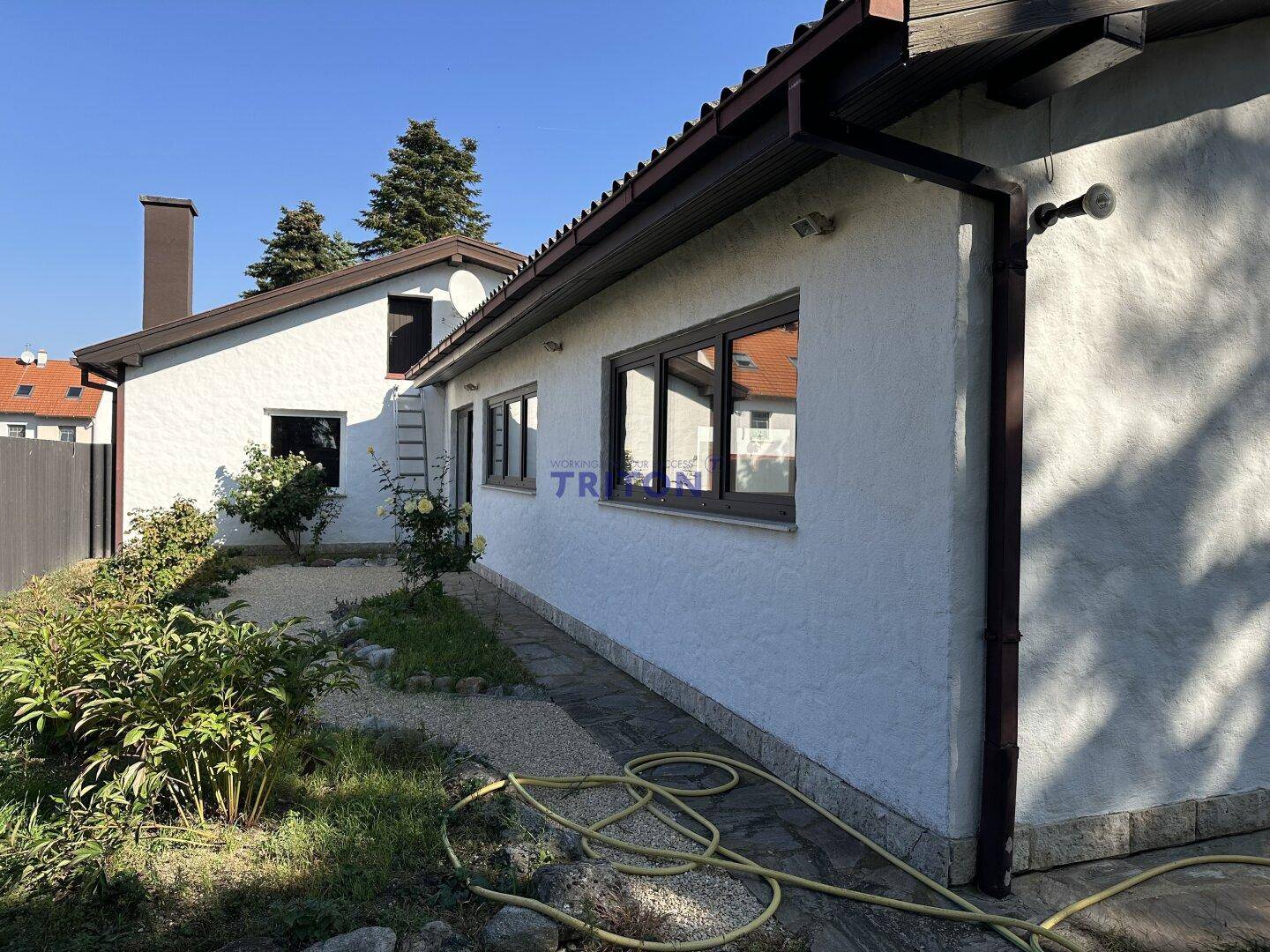 Seitenansicht des Hauses mit kleiner Gartenanlage, Blumenbeeten und Schlauch.